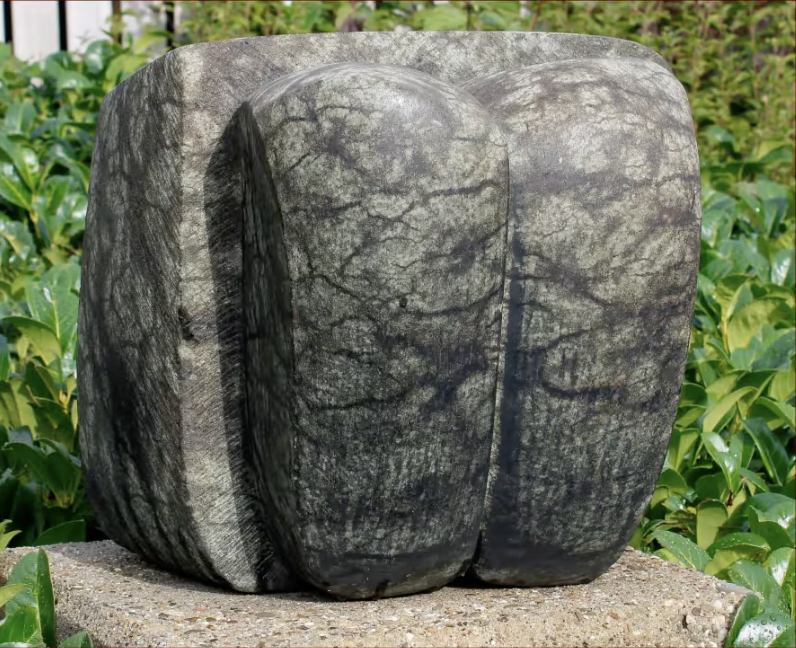 Abstracte stenen sculptuur met twee ronde, verticale vormen naast elkaar, geplaatst op een betonnen sokkel, omgeven door een groene heg met glanzende bladeren