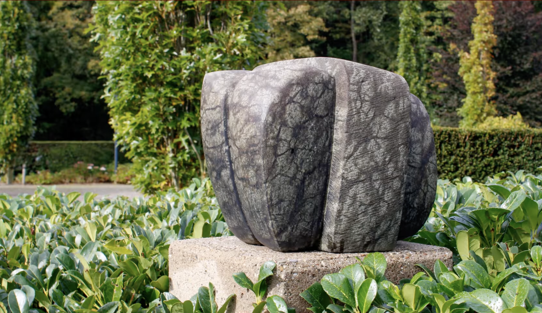 Abstracte stenen sculptuur met twee ronde, verticale vormen naast elkaar, geplaatst op een betonnen sokkel, omgeven door een groene heg met glanzende bladeren
