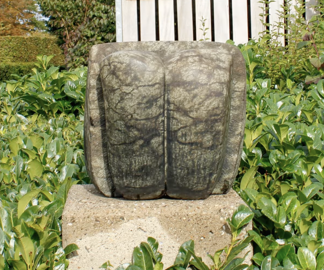 Abstracte stenen sculptuur met twee ronde, verticale vormen naast elkaar, geplaatst op een betonnen sokkel, omgeven door een groene heg met glanzende bladeren