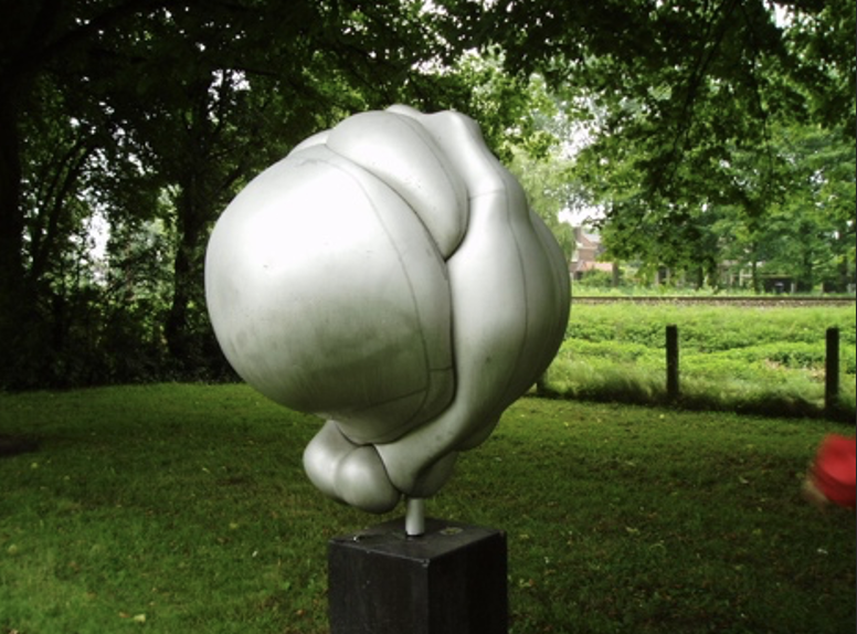 Een abstracte, witte sculptuur van organische, vloeiende vormen opgesteld in een groene buitenruimte met bomen op de achtergrond.