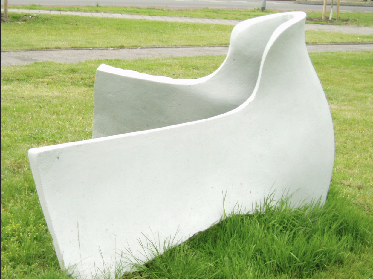Wit abstract kunstwerk in de vorm van gebogen, golvende platen op een grasveld naast een fietspad, met een weg op de achtergrond