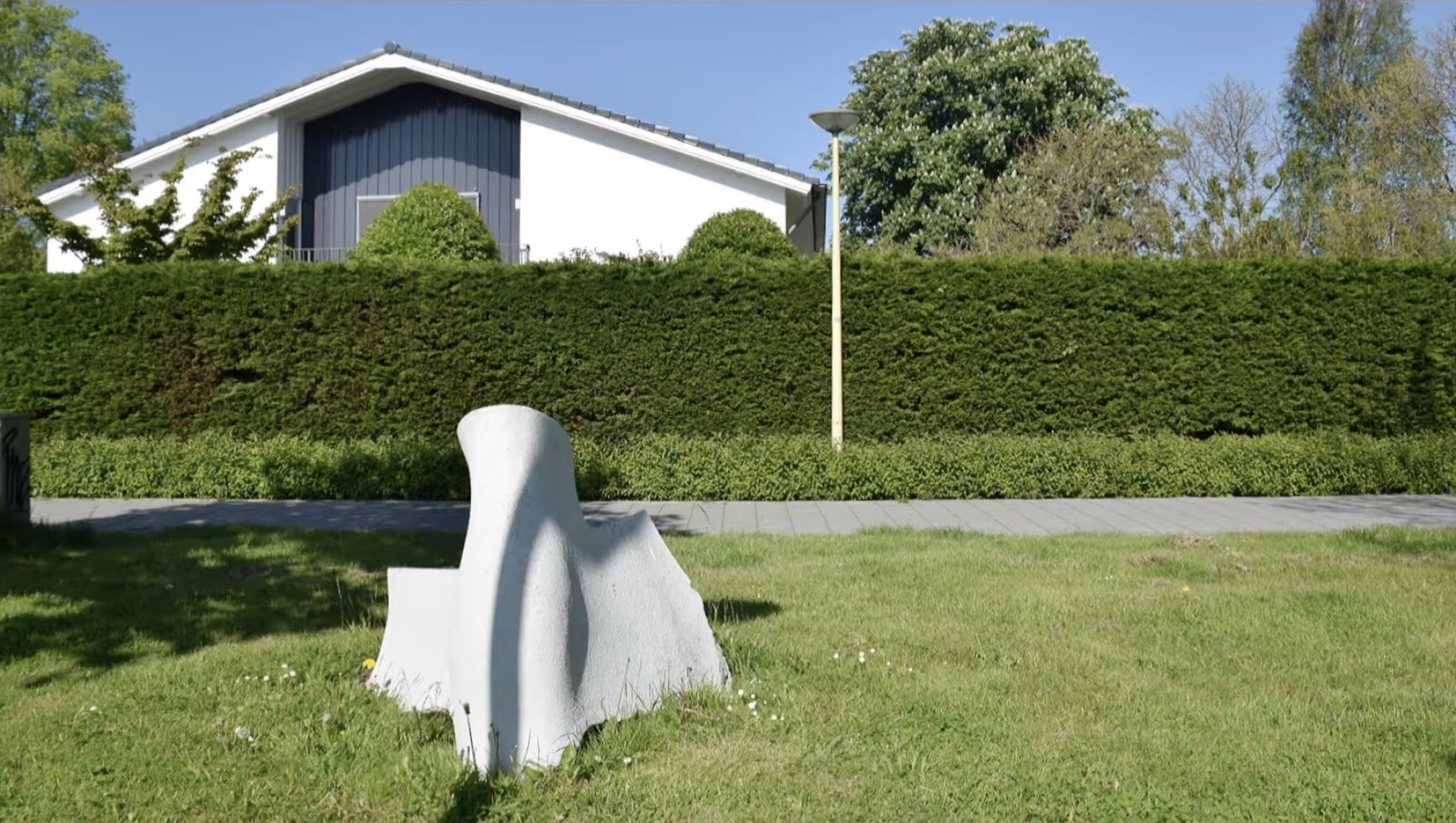 Wit abstract kunstwerk in de vorm van gebogen, golvende platen op een grasveld naast een fietspad, met een weg op de achtergrond