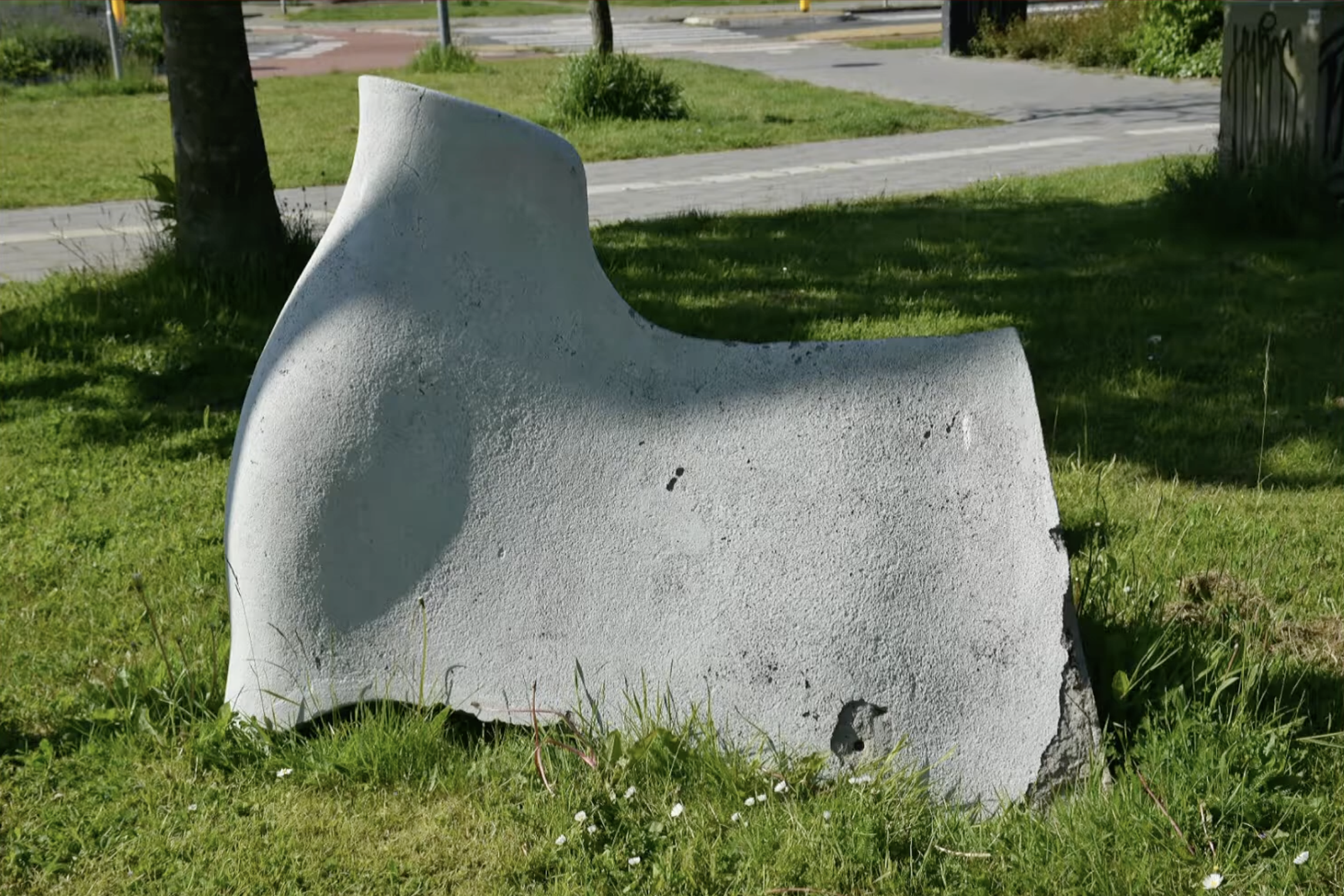 Wit abstract kunstwerk in de vorm van gebogen, golvende platen op een grasveld naast een fietspad, met een weg op de achtergrond