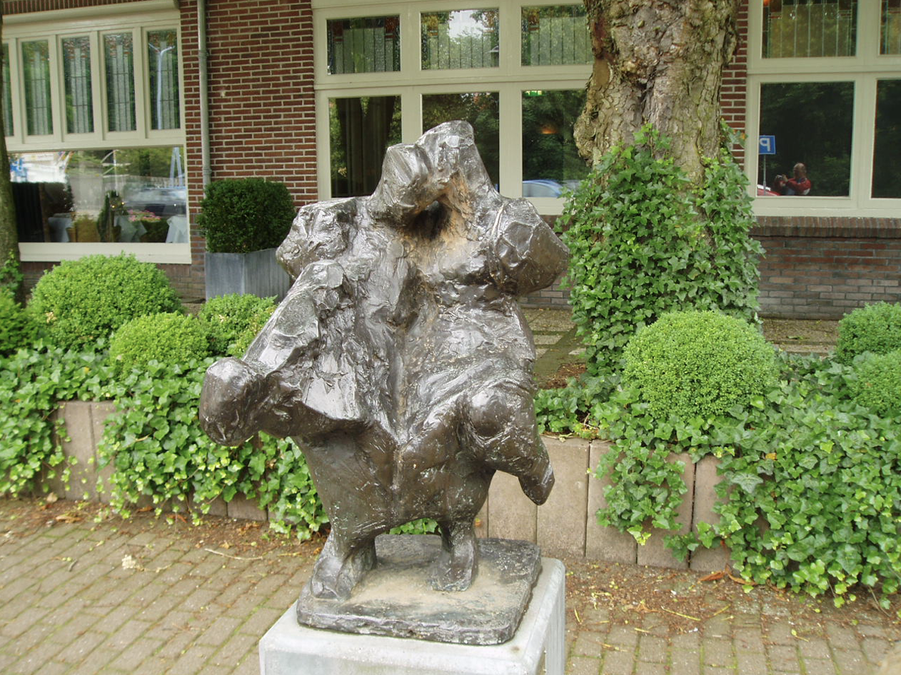 Bronzen abstract beeld met een ruw, gescheurd oppervlak op een stenen sokkel, voor een bakstenen gebouw met klimop en een boom.