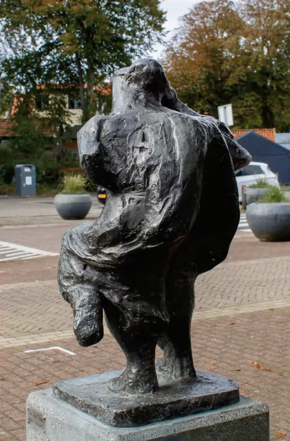Bronzen abstract beeld met een ruw, gescheurd oppervlak op een stenen sokkel, voor een bakstenen gebouw met klimop en een boom.