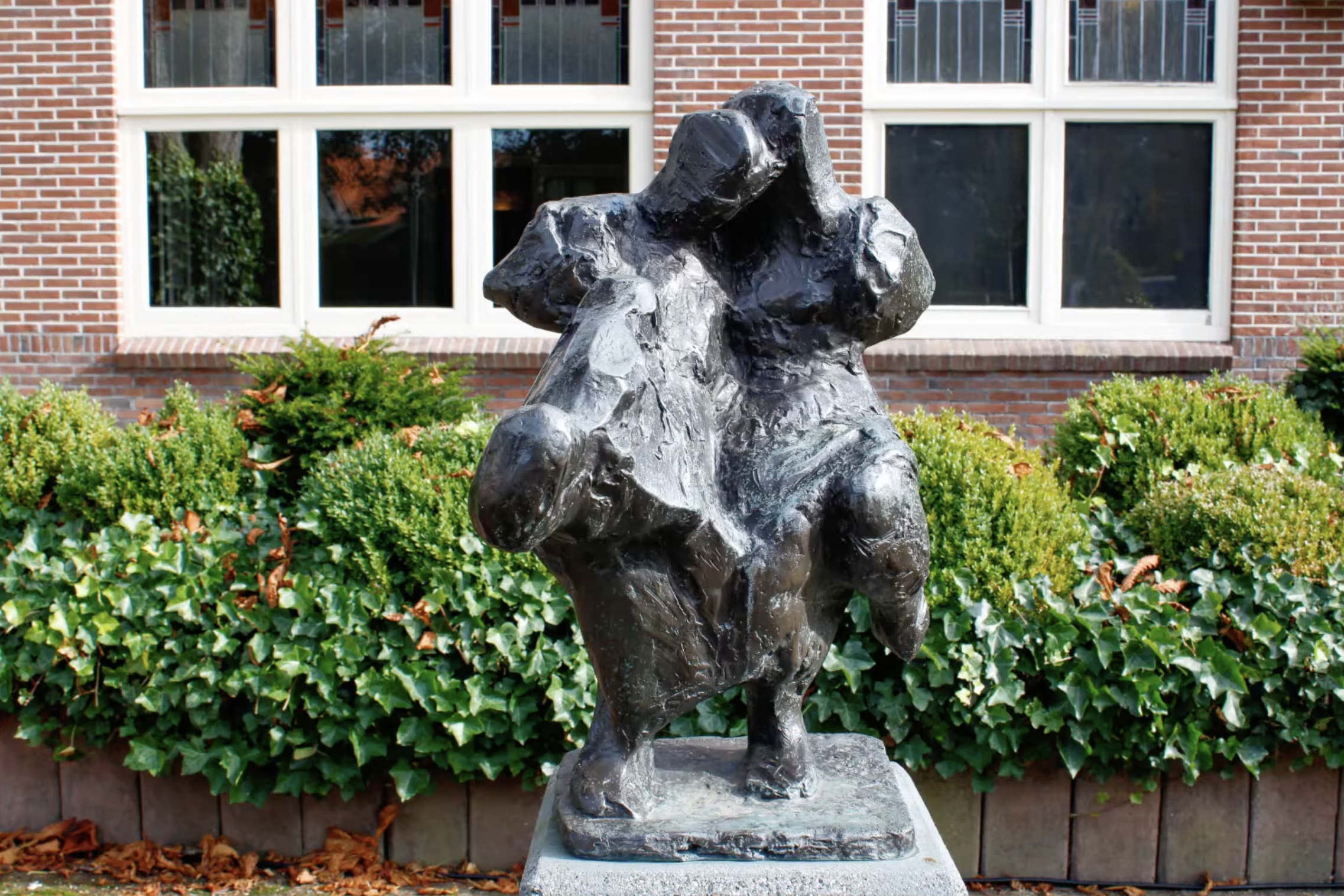 Bronzen abstract beeld met een ruw, gescheurd oppervlak op een stenen sokkel, voor een bakstenen gebouw met klimop en een boom.