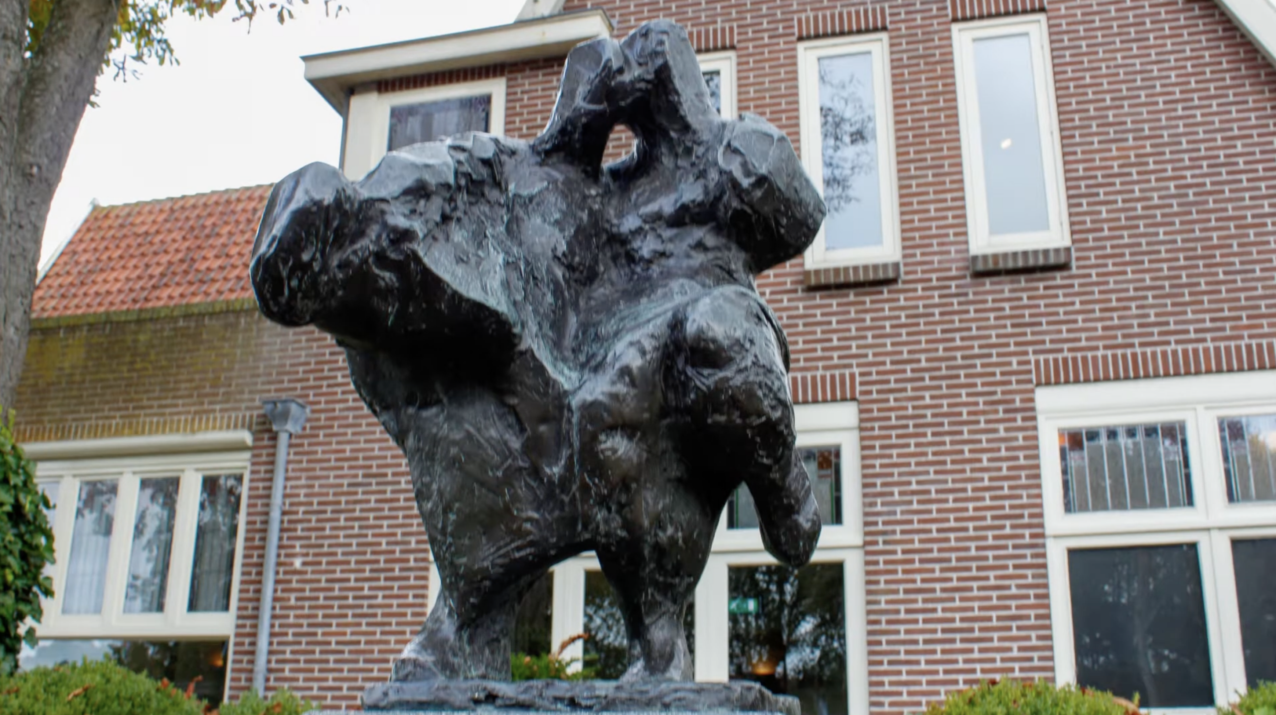 Bronzen abstract beeld met een ruw, gescheurd oppervlak op een stenen sokkel, voor een bakstenen gebouw met klimop en een boom.