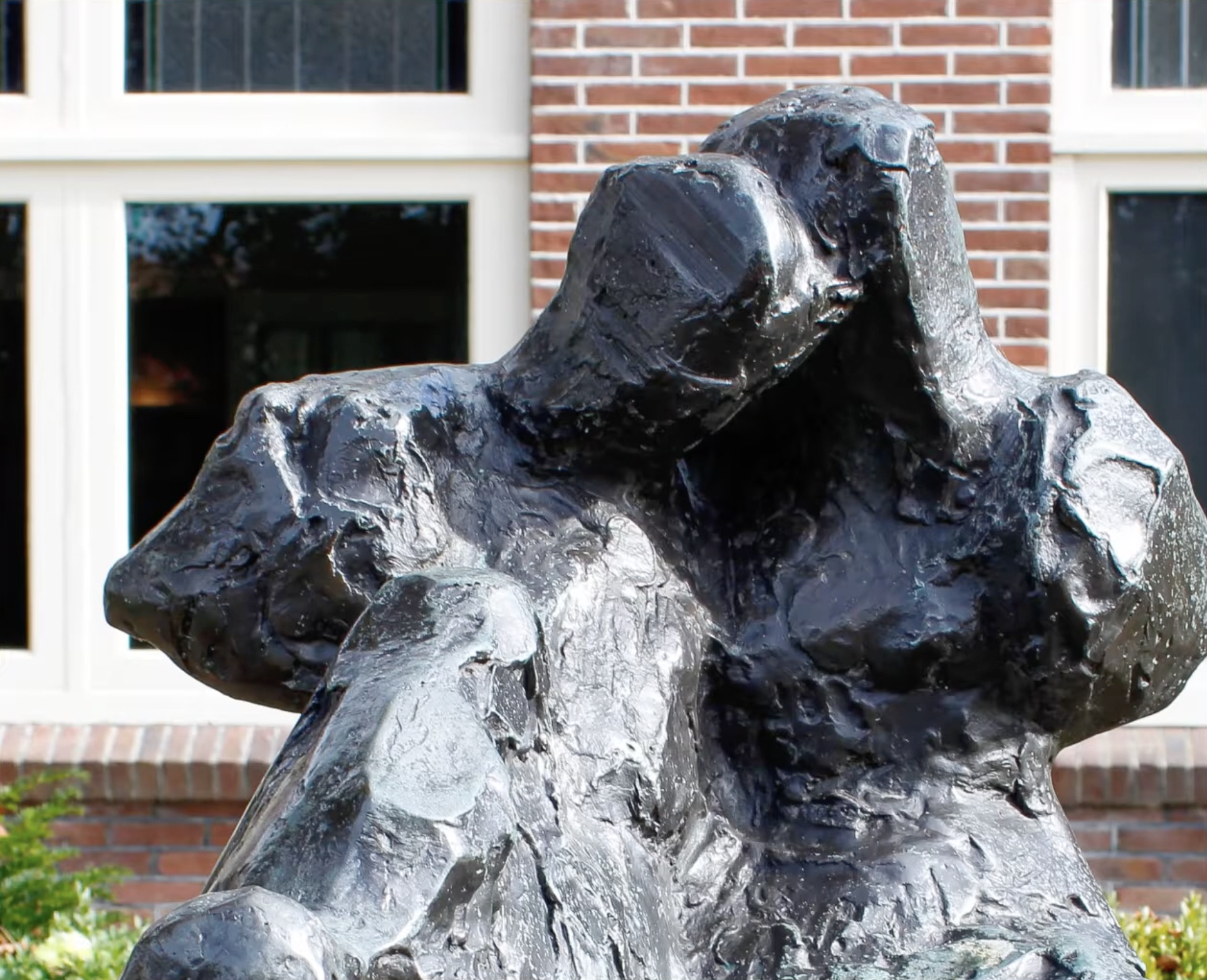 Bronzen abstract beeld met een ruw, gescheurd oppervlak op een stenen sokkel, voor een bakstenen gebouw met klimop en een boom.