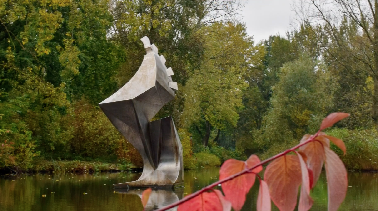 Metalen sculptuur met gedraaide, bladachtige vormen staat in een vijver, omringd door groen en bomen