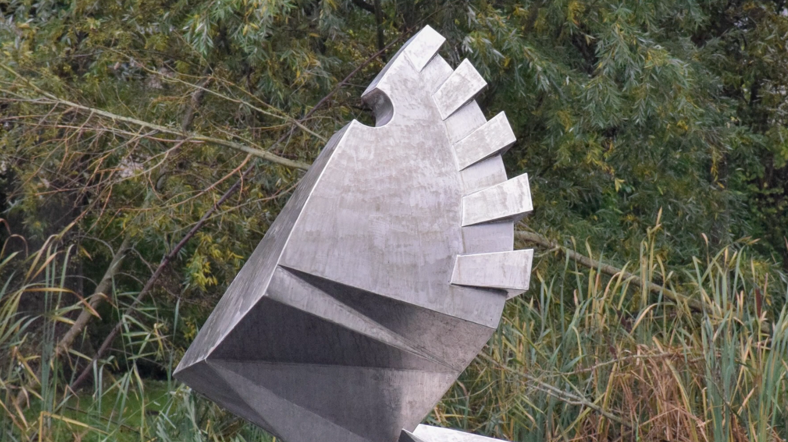 Metalen sculptuur met gedraaide, bladachtige vormen staat in een vijver, omringd door groen en bomen