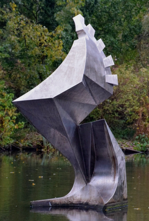 Metalen sculptuur met gedraaide, bladachtige vormen staat in een vijver, omringd door groen en bomen
