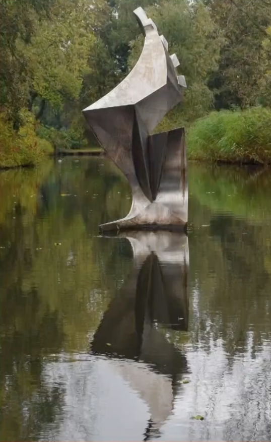 Metalen sculptuur met gedraaide, bladachtige vormen staat in een vijver, omringd door groen en bomen