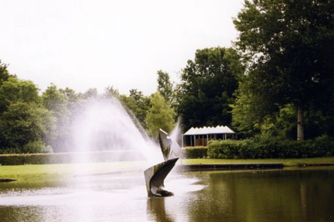 Metalen sculptuur met gedraaide, bladachtige vormen staat in een vijver, omringd door groen en bomen