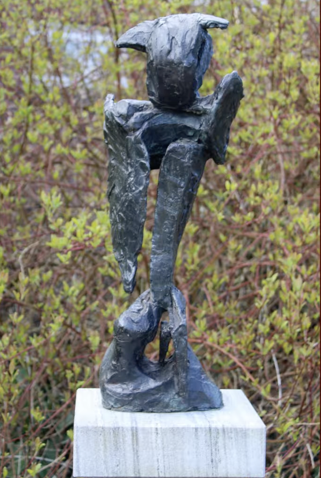 Een donker, ruw getextureerd bronzen beeld met een verticale, abstracte vorm op een stenen sokkel, met een groene struikenrand