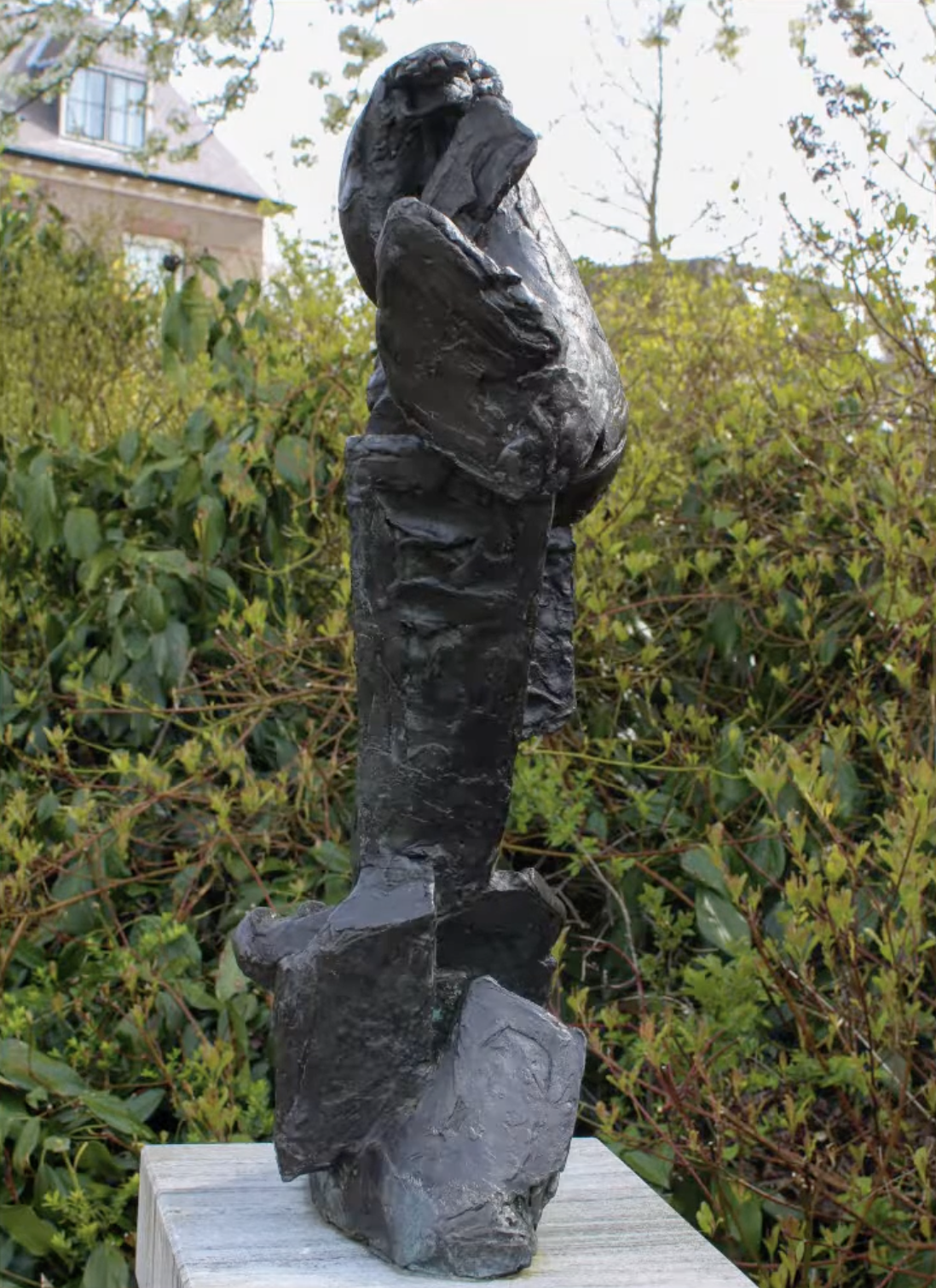 Een donker, ruw getextureerd bronzen beeld met een verticale, abstracte vorm op een stenen sokkel, met een groene struikenrand