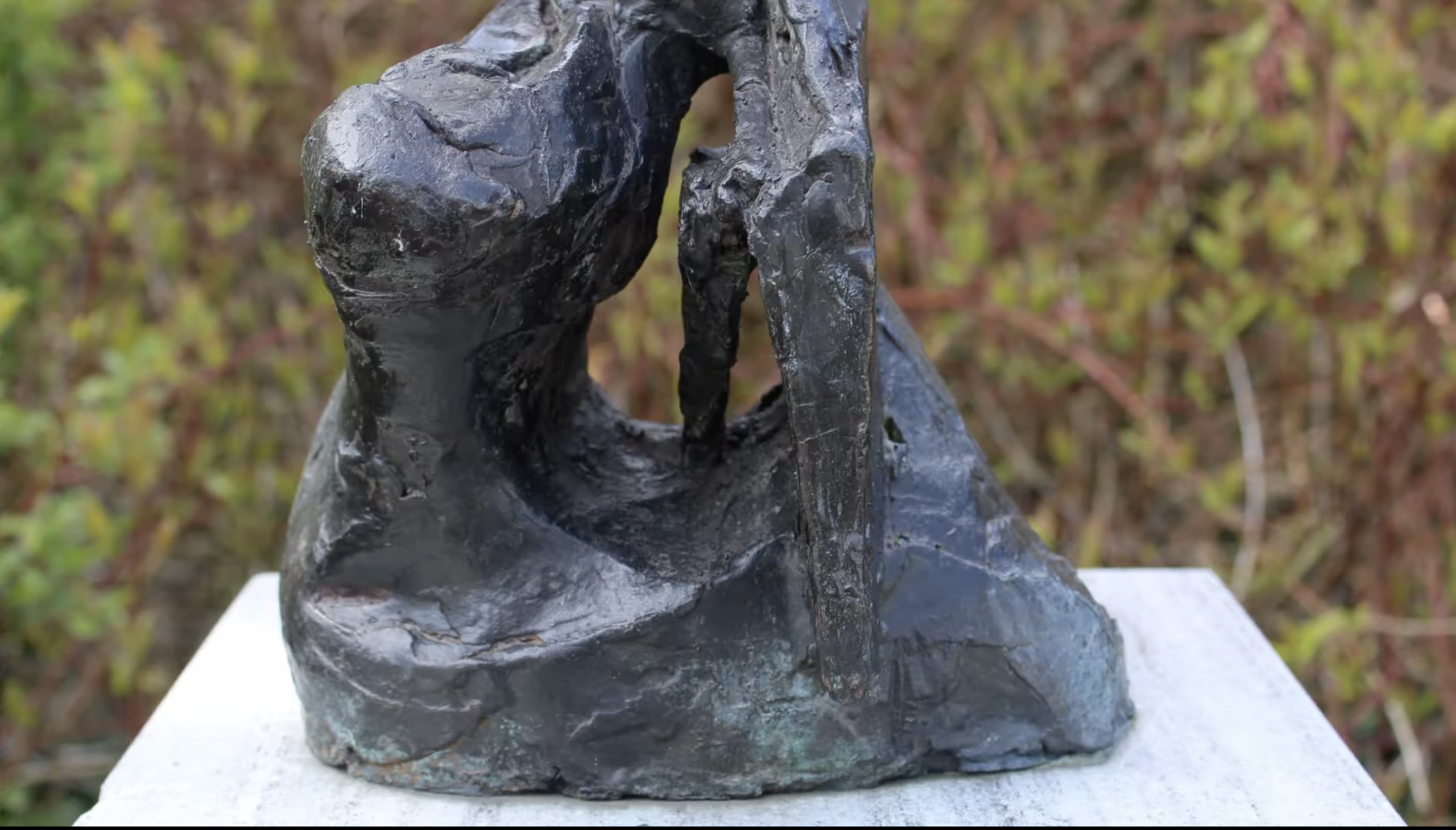 Een donker, ruw getextureerd bronzen beeld met een verticale, abstracte vorm op een stenen sokkel, met een groene struikenrand