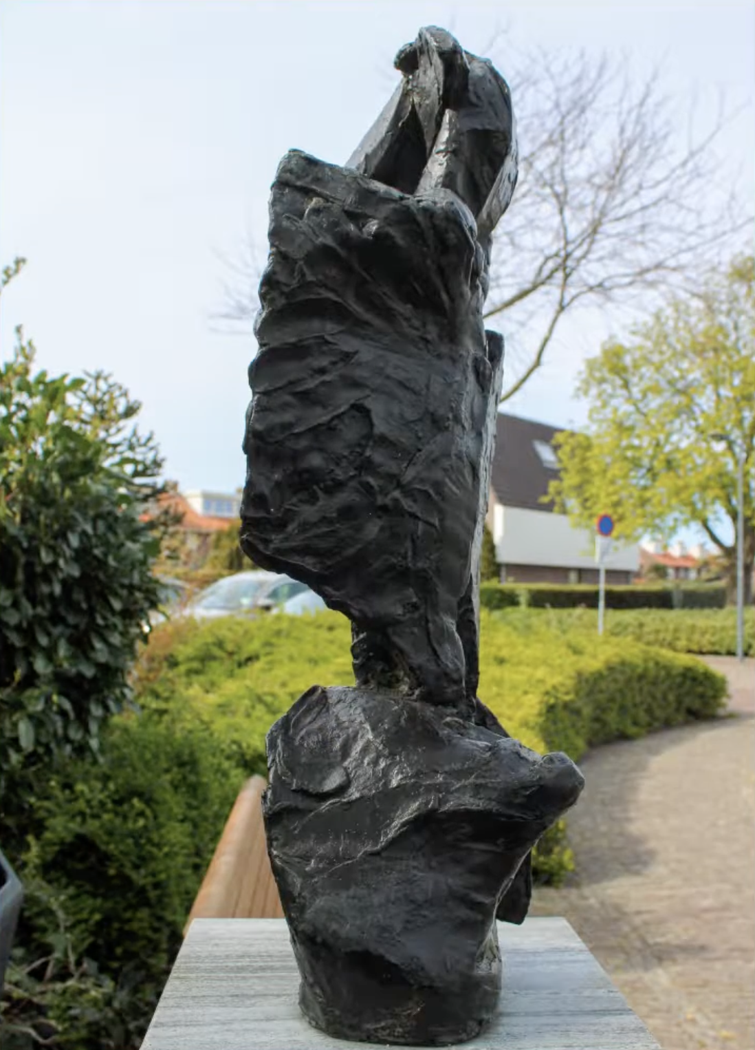 Een donker, ruw getextureerd bronzen beeld met een verticale, abstracte vorm op een stenen sokkel, met een groene struikenrand
