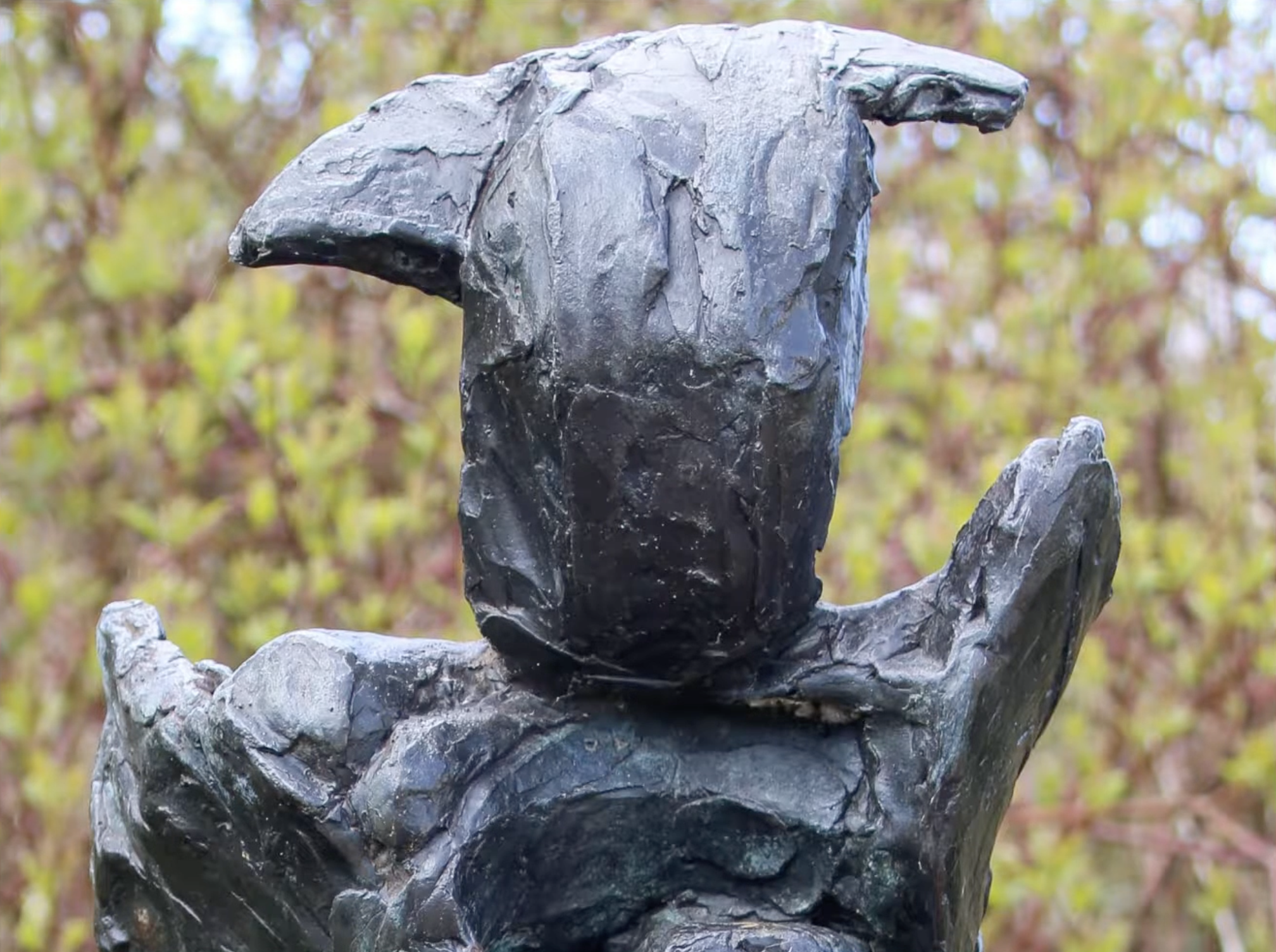 Een donker, ruw getextureerd bronzen beeld met een verticale, abstracte vorm op een stenen sokkel, met een groene struikenrand