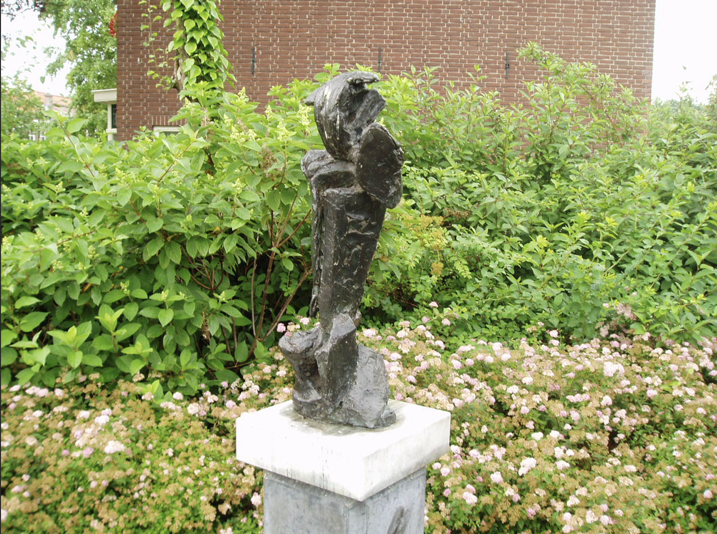 Een donker, ruw getextureerd bronzen beeld met een verticale, abstracte vorm op een stenen sokkel, met een groene struikenrand