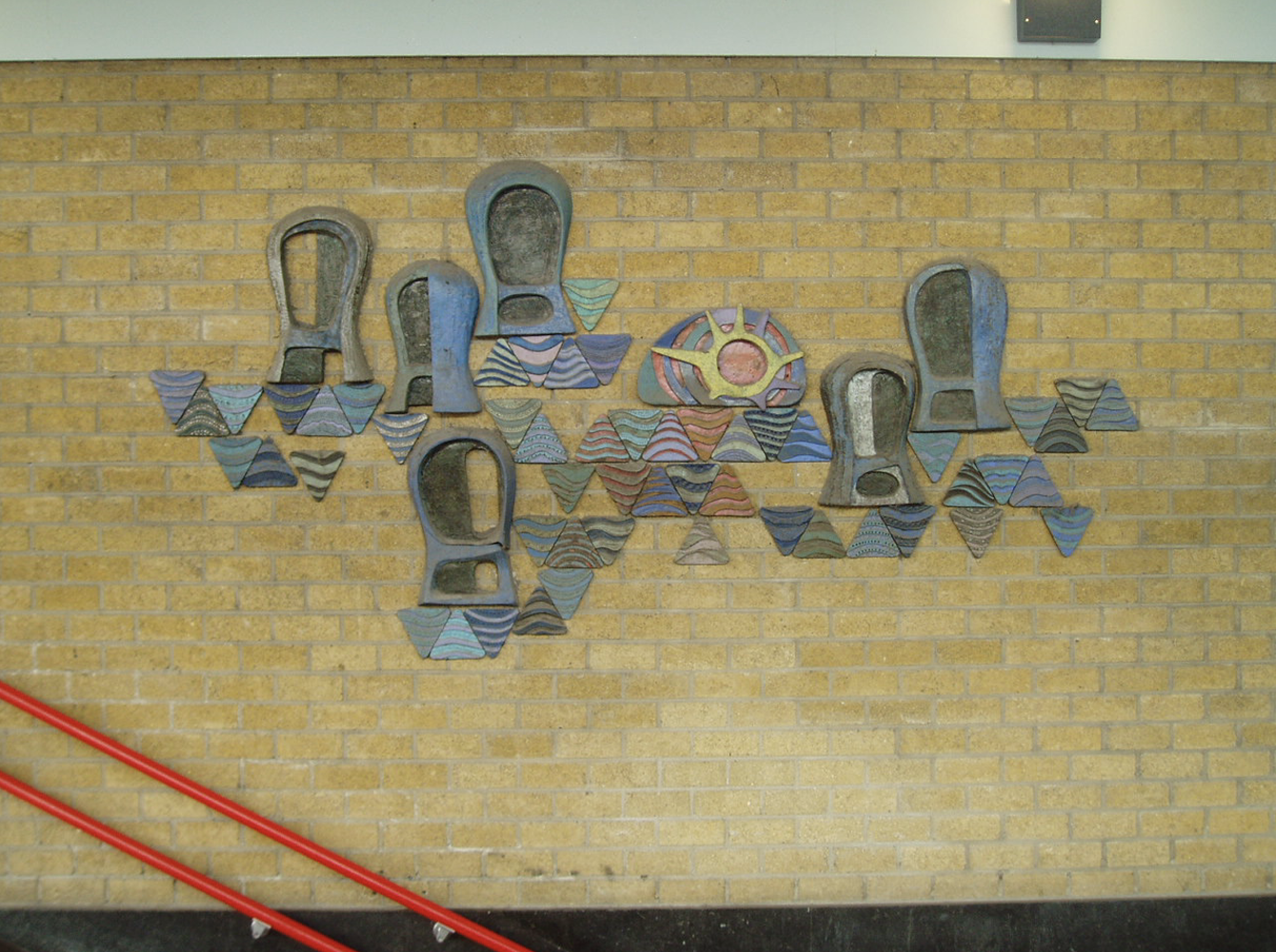 Een keramisch wandkunstwerk op een gele bakstenen muur, met abstracte, kapvormige figuren en gekleurde driehoekige elementen in blauw-, grijs- en aardetinten, met een rode trapleuning op de voorgrond
