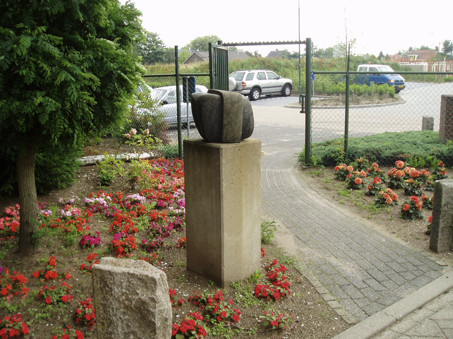 Een stenen beeld op een betonnen sokkel in een bloemperk met rode en roze bloemen, bij een hek en parkeerplaats met auto's op de achtergrond.