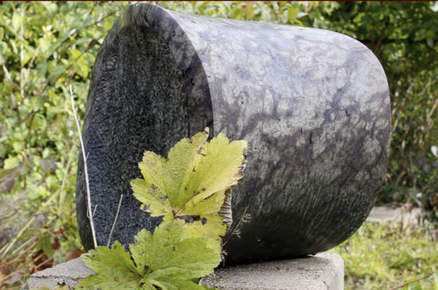Een cilindervormig stenen beeld, liggend op een betonnen sokkel, omringd door groene struiken en coniferen.