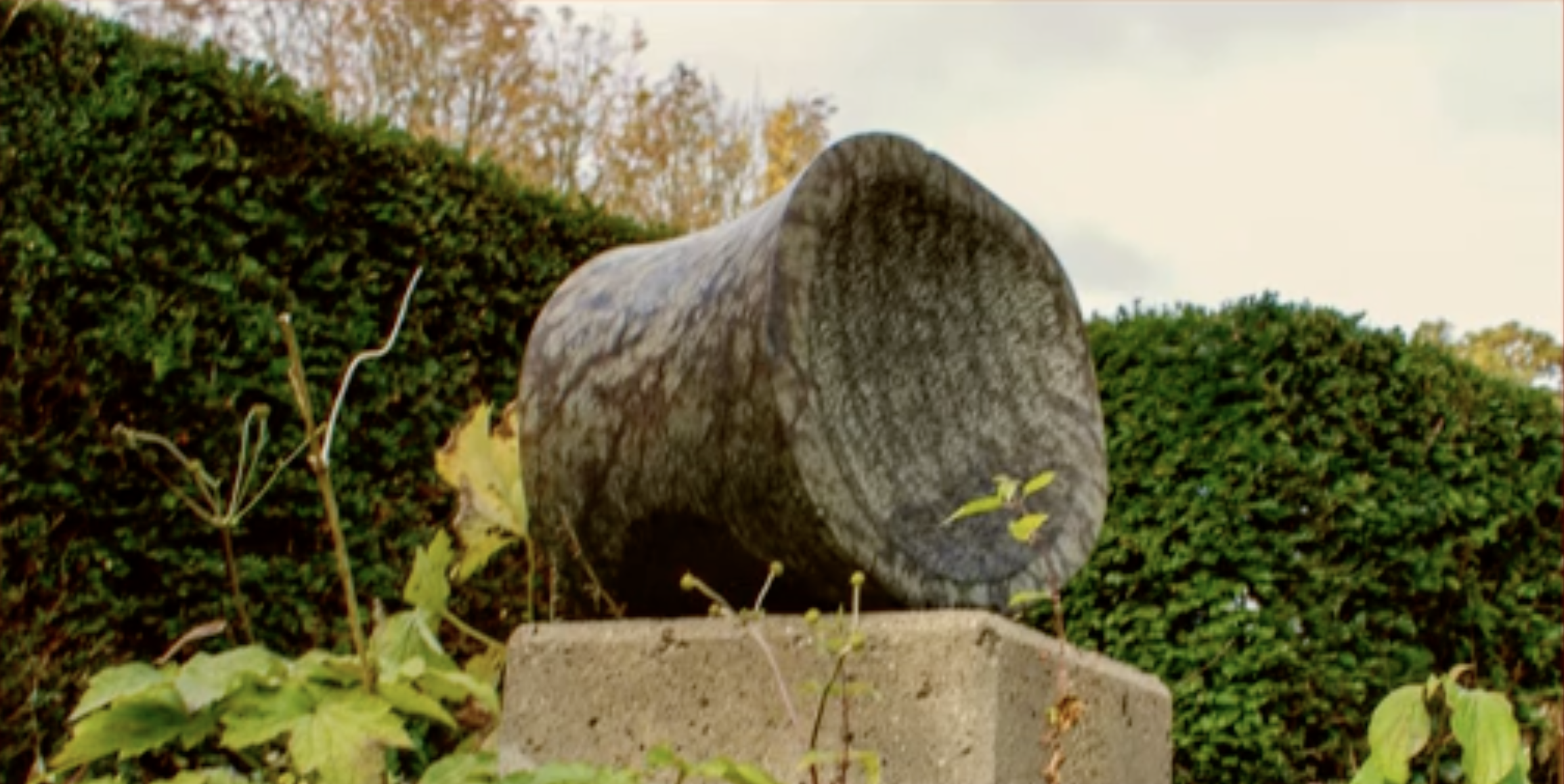 Een cilindervormig stenen beeld, liggend op een betonnen sokkel, omringd door groene struiken en coniferen.