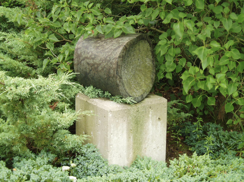 Een cilindervormig stenen beeld, liggend op een betonnen sokkel, omringd door groene struiken en coniferen.