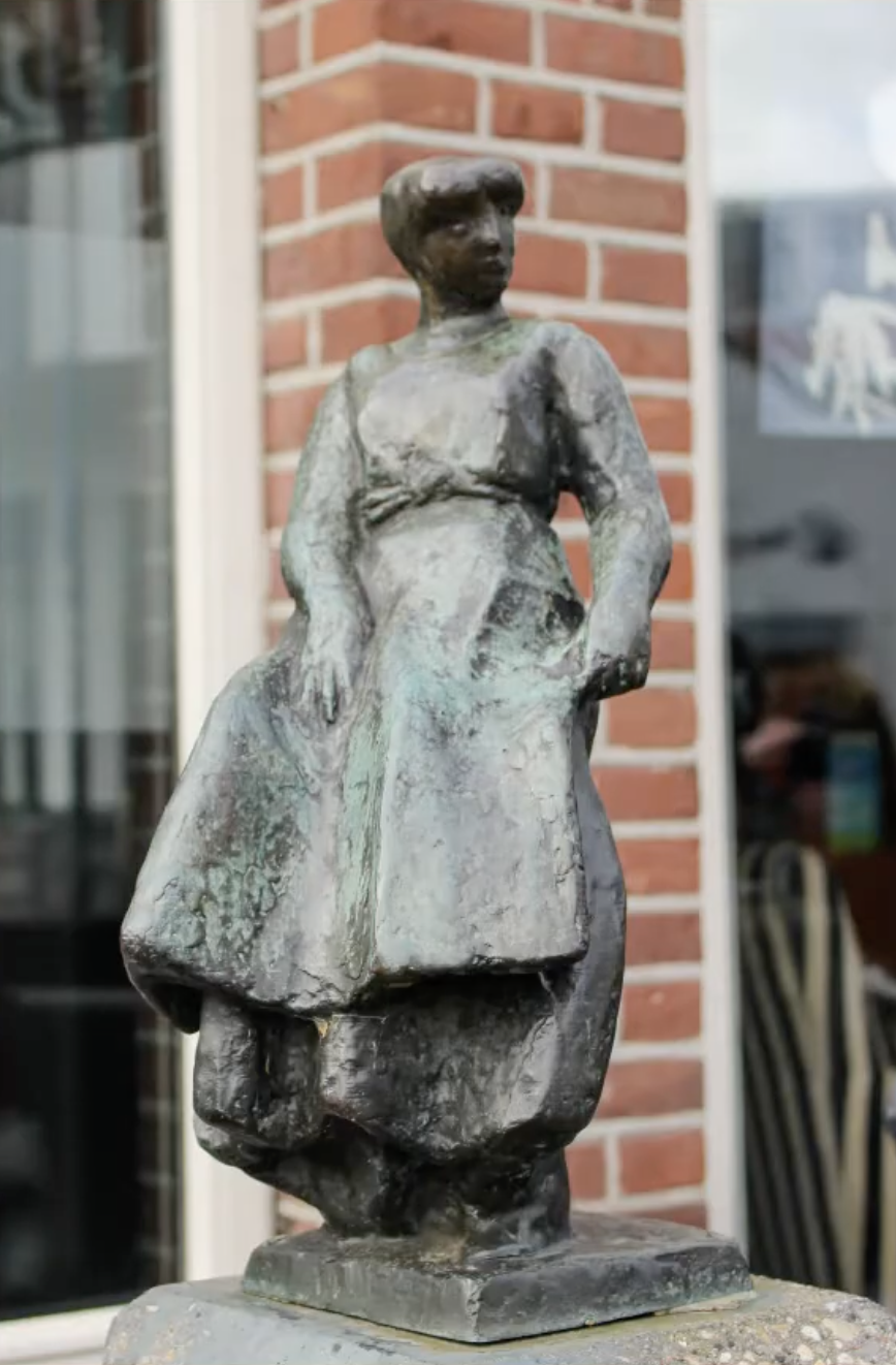 Een klein bronzen beeldje van een vrouwenfiguur in traditionele klederdracht, geplaatst op een hoge stenen sokkel op een stoep van klinkers, voor een winkel met een open-bordje in het raam.