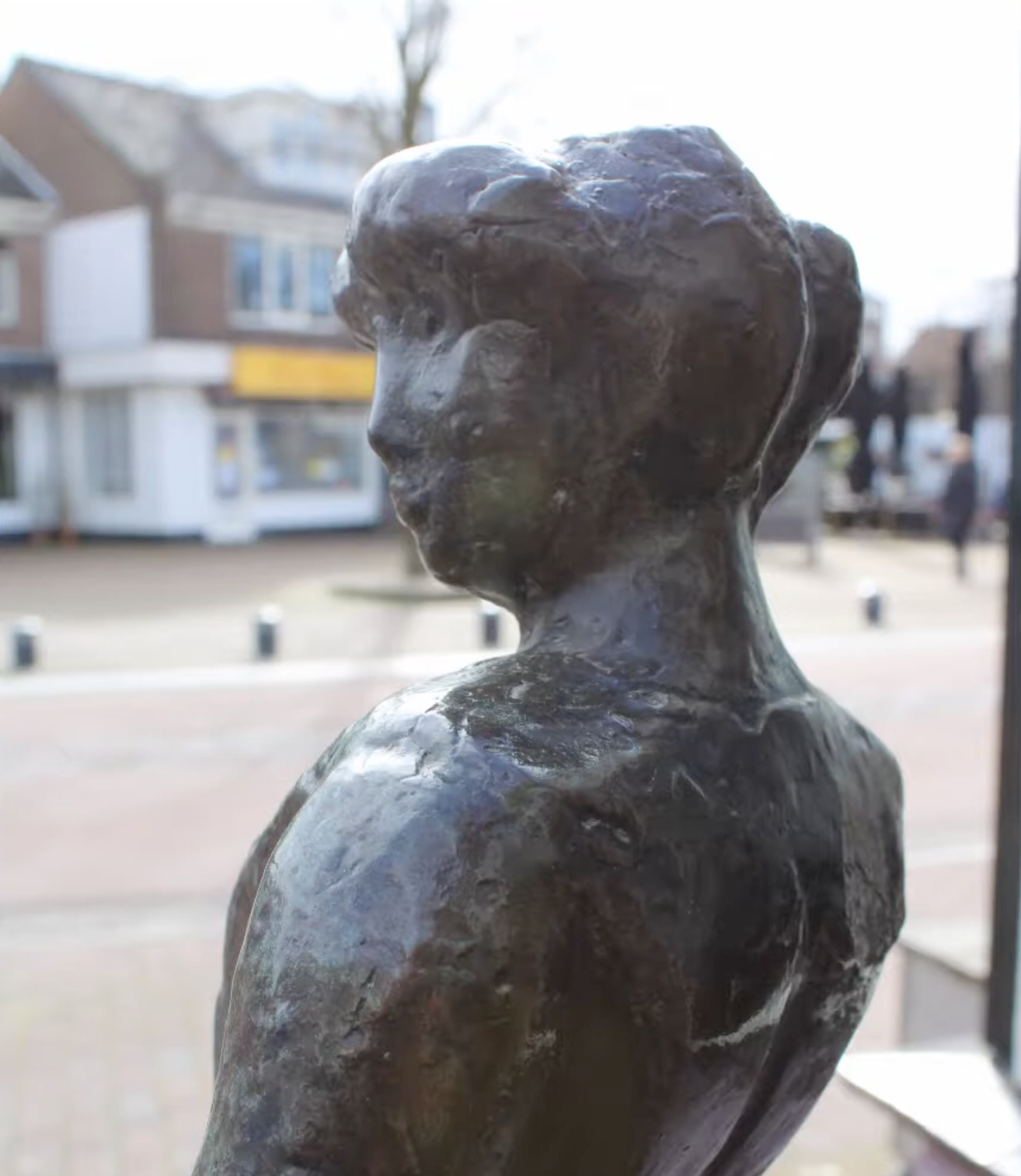 Een klein bronzen beeldje van een vrouwenfiguur in traditionele klederdracht, geplaatst op een hoge stenen sokkel op een stoep van klinkers, voor een winkel met een open-bordje in het raam.