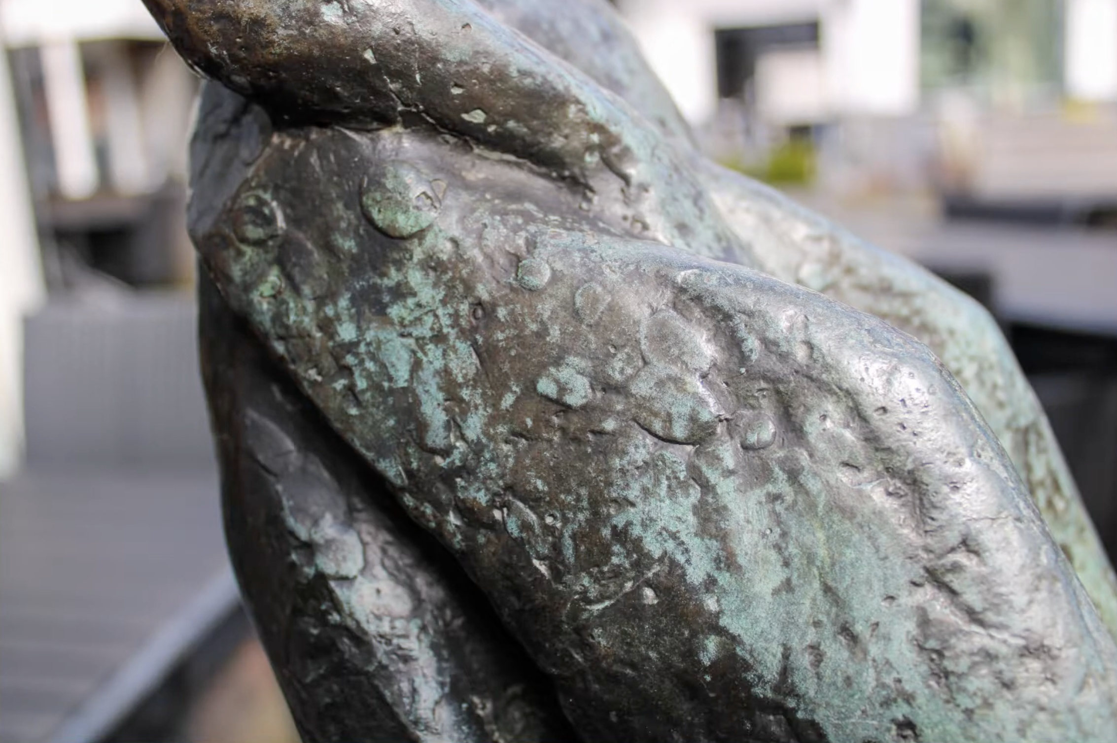 Een klein bronzen beeldje van een vrouwenfiguur in traditionele klederdracht, geplaatst op een hoge stenen sokkel op een stoep van klinkers, voor een winkel met een open-bordje in het raam.