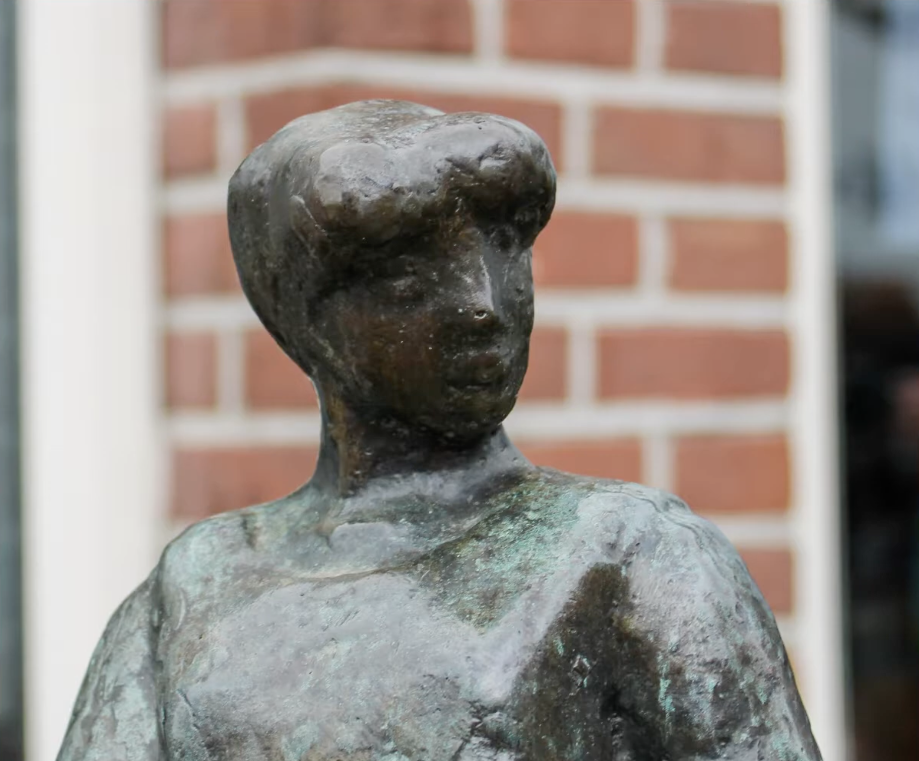 Een klein bronzen beeldje van een vrouwenfiguur in traditionele klederdracht, geplaatst op een hoge stenen sokkel op een stoep van klinkers, voor een winkel met een open-bordje in het raam.