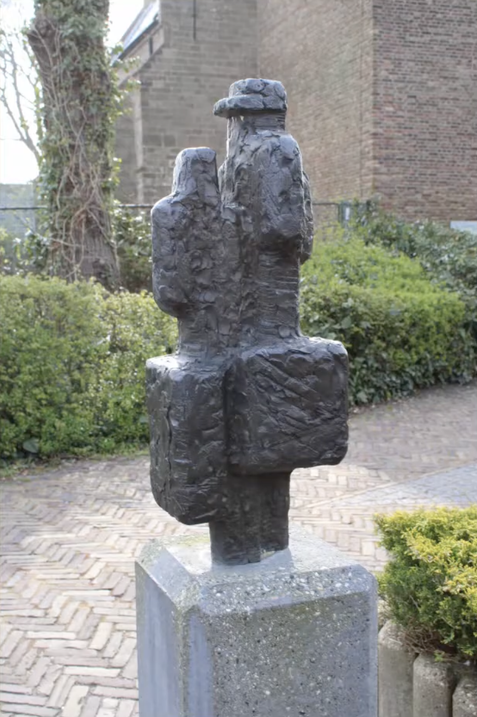 Een donker bronzen beeldje van twee menselijke figuren naast elkaar, geplaatst op een met mos begroeide stenen sokkel, omgeven door weelderig groen struikgewas en een klinkerpad op de achtergrond.