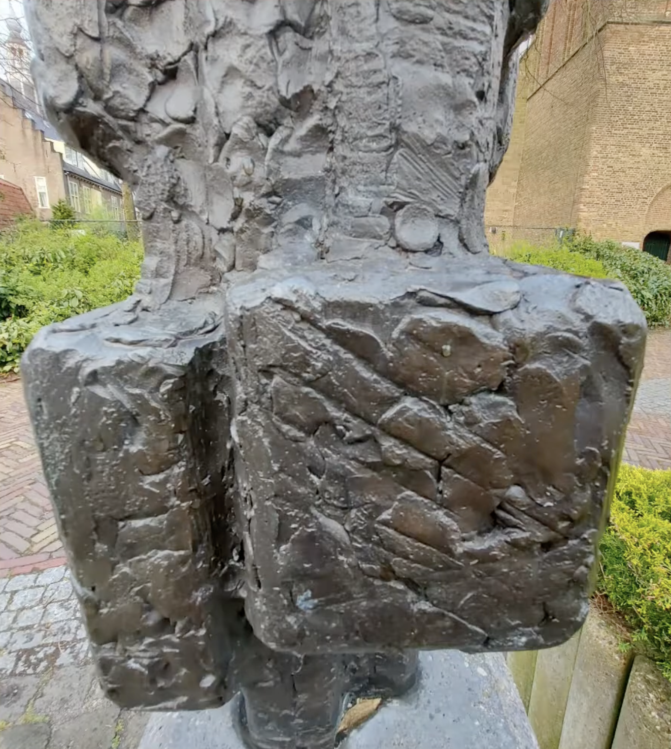 Een donker bronzen beeldje van twee menselijke figuren naast elkaar, geplaatst op een met mos begroeide stenen sokkel, omgeven door weelderig groen struikgewas en een klinkerpad op de achtergrond.