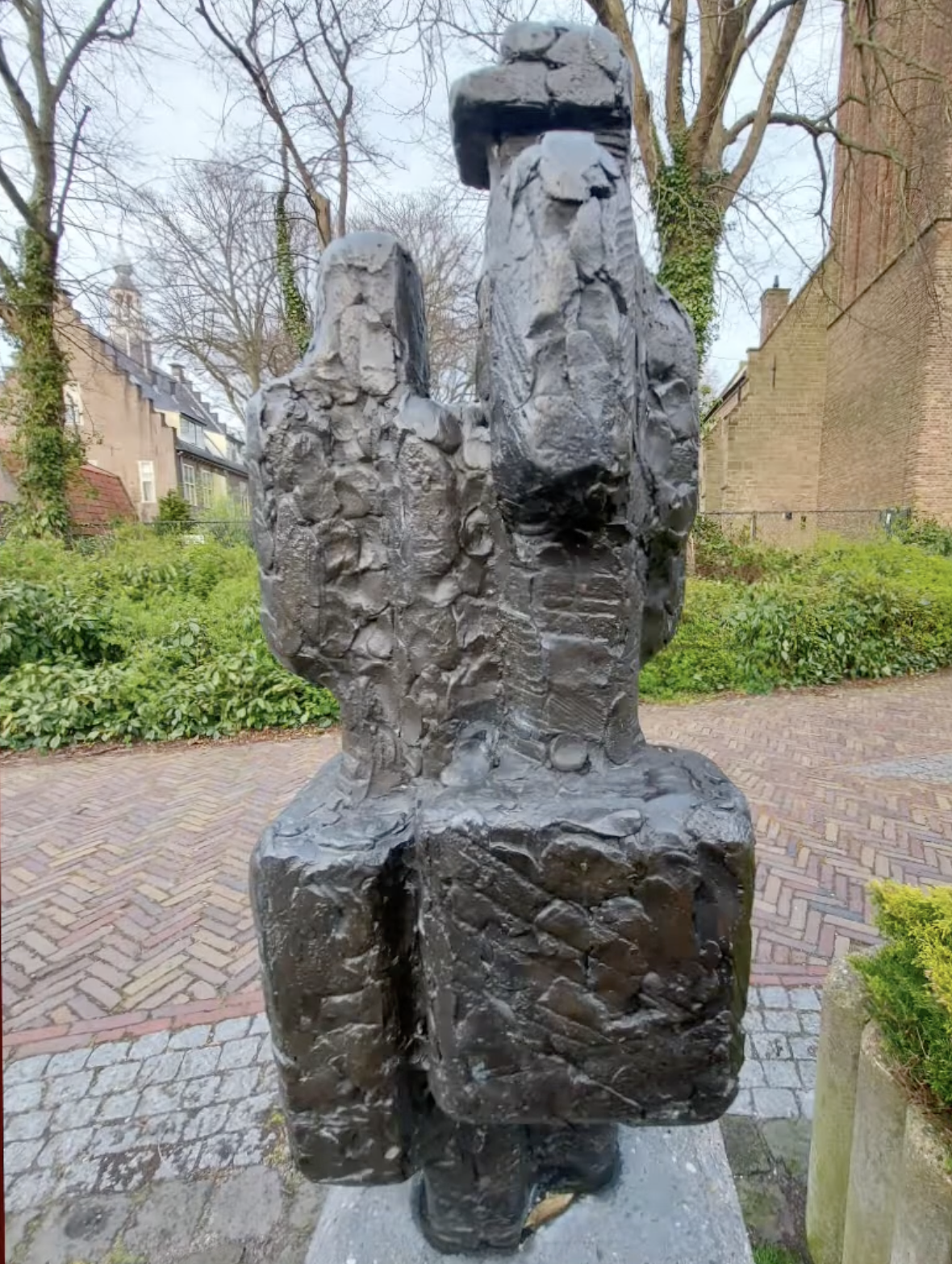 Een donker bronzen beeldje van twee menselijke figuren naast elkaar, geplaatst op een met mos begroeide stenen sokkel, omgeven door weelderig groen struikgewas en een klinkerpad op de achtergrond.