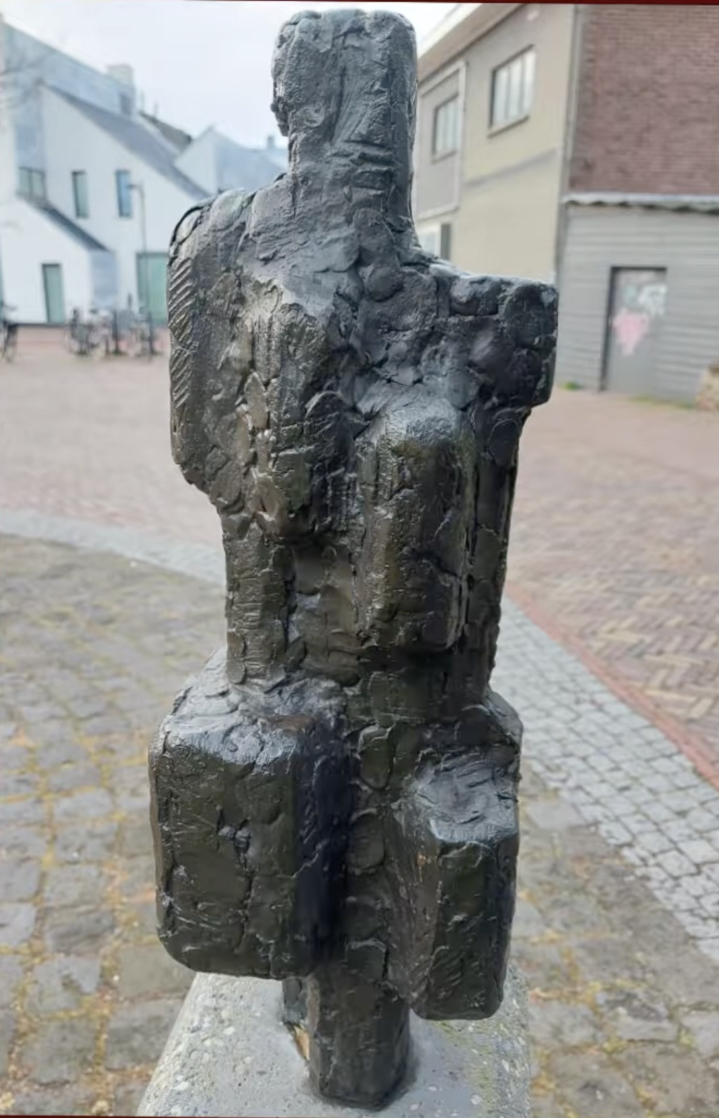 Een donker bronzen beeldje van twee menselijke figuren naast elkaar, geplaatst op een met mos begroeide stenen sokkel, omgeven door weelderig groen struikgewas en een klinkerpad op de achtergrond.