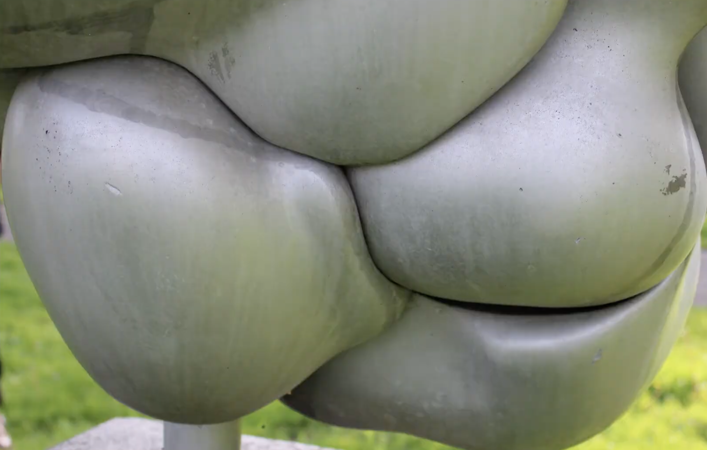 Een abstracte, witte sculptuur van organische, vloeiende vormen opgesteld in een groene buitenruimte met bomen op de achtergrond.