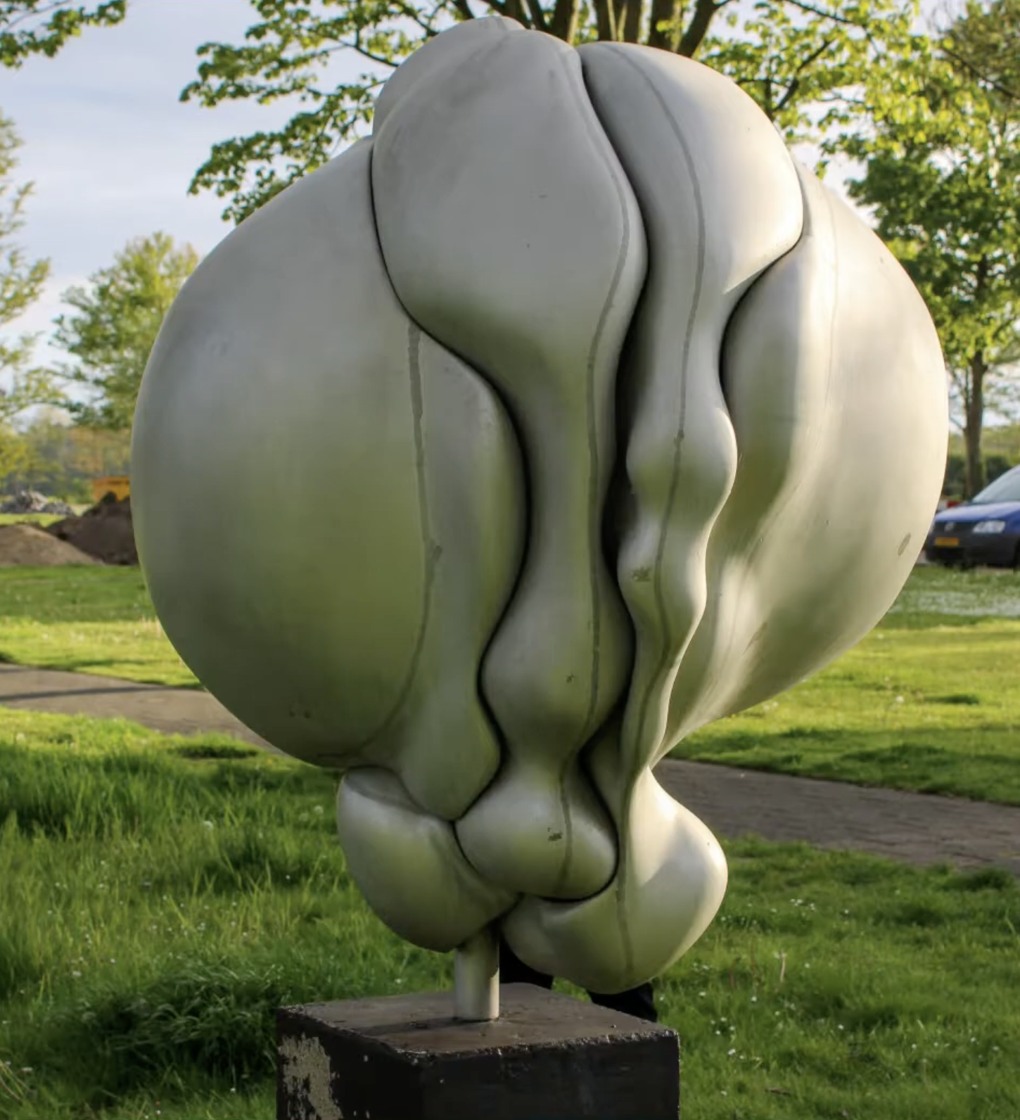 Een abstracte, witte sculptuur van organische, vloeiende vormen opgesteld in een groene buitenruimte met bomen op de achtergrond.