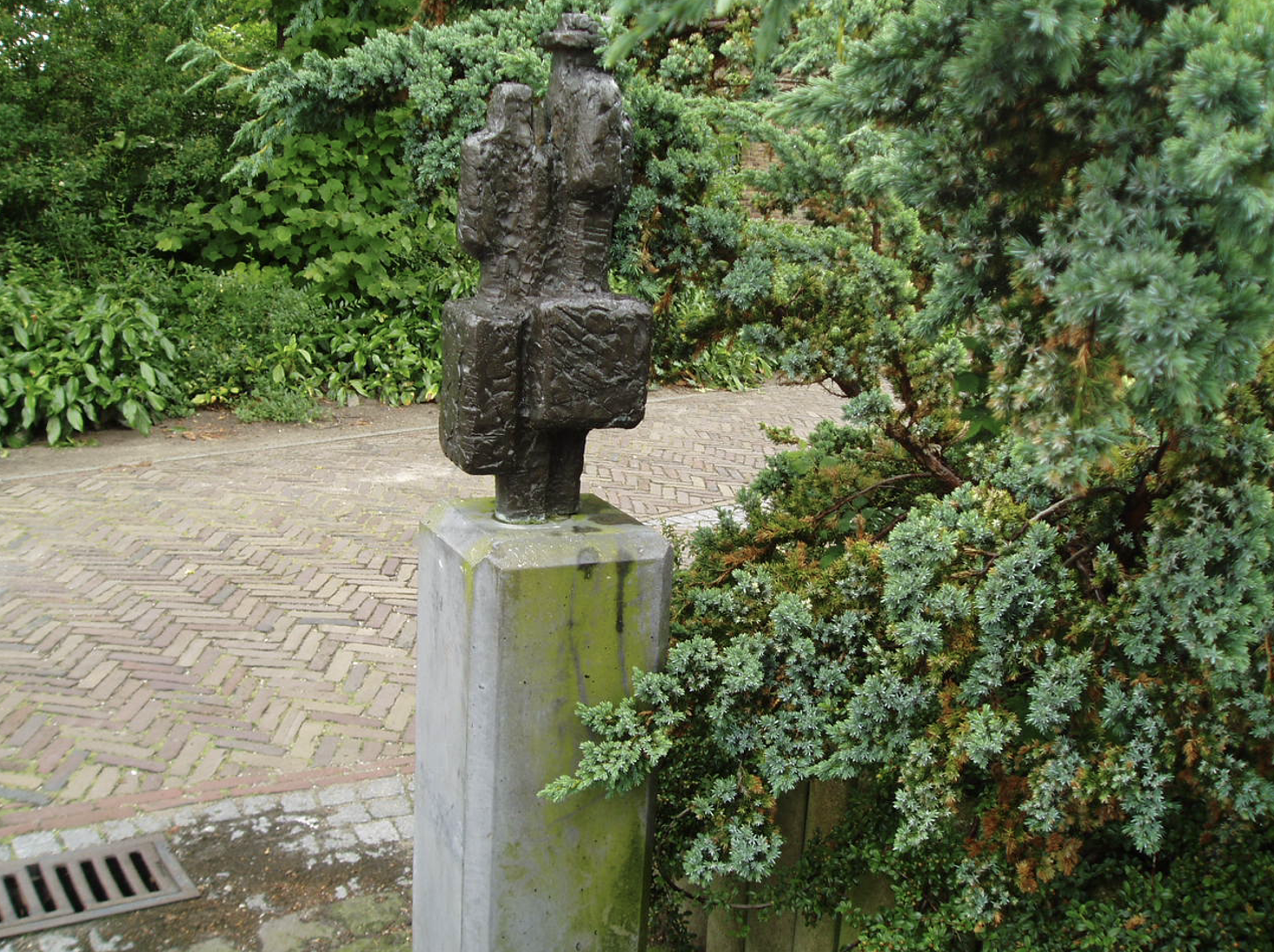 Een donker bronzen beeldje van twee menselijke figuren naast elkaar, geplaatst op een met mos begroeide stenen sokkel, omgeven door weelderig groen struikgewas en een klinkerpad op de achtergrond.