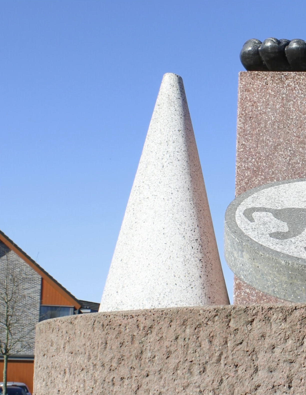Abstracte sculptuur van terrazzo en brons op drie pijlers, met een voet, een kegel en een reliëfschijf, in een parkachtige omgeving.