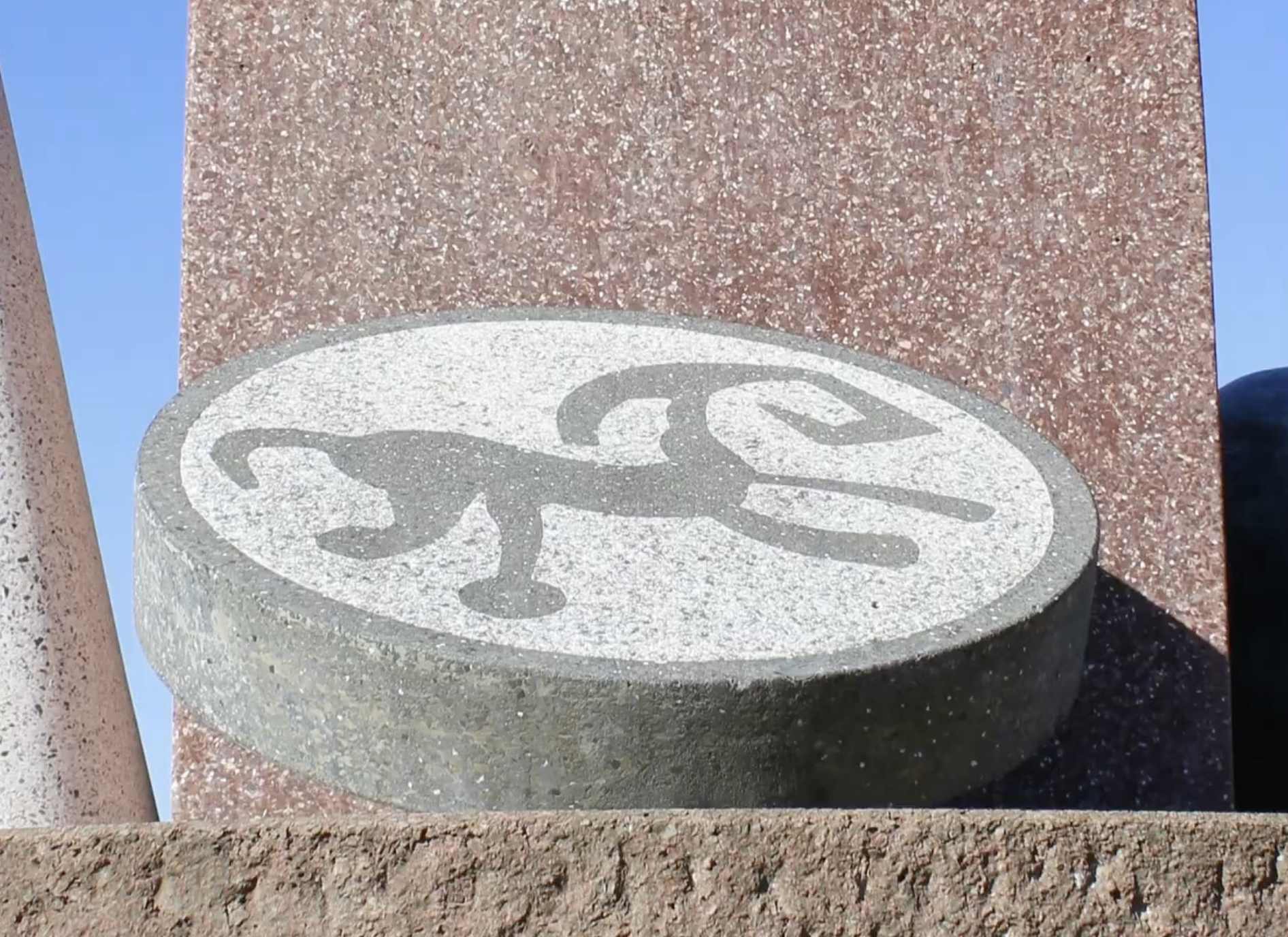 Abstracte sculptuur van terrazzo en brons op drie pijlers, met een voet, een kegel en een reliëfschijf, in een parkachtige omgeving.