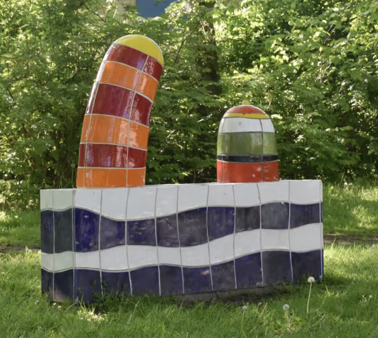 Kleurrijke keramische sculptuur met blauwe en witte golfmotieven op de sokkel en twee felgekleurde torenvormige elementen in rood, oranje en geel, opgesteld in een groenstrook langs een woonstraat.