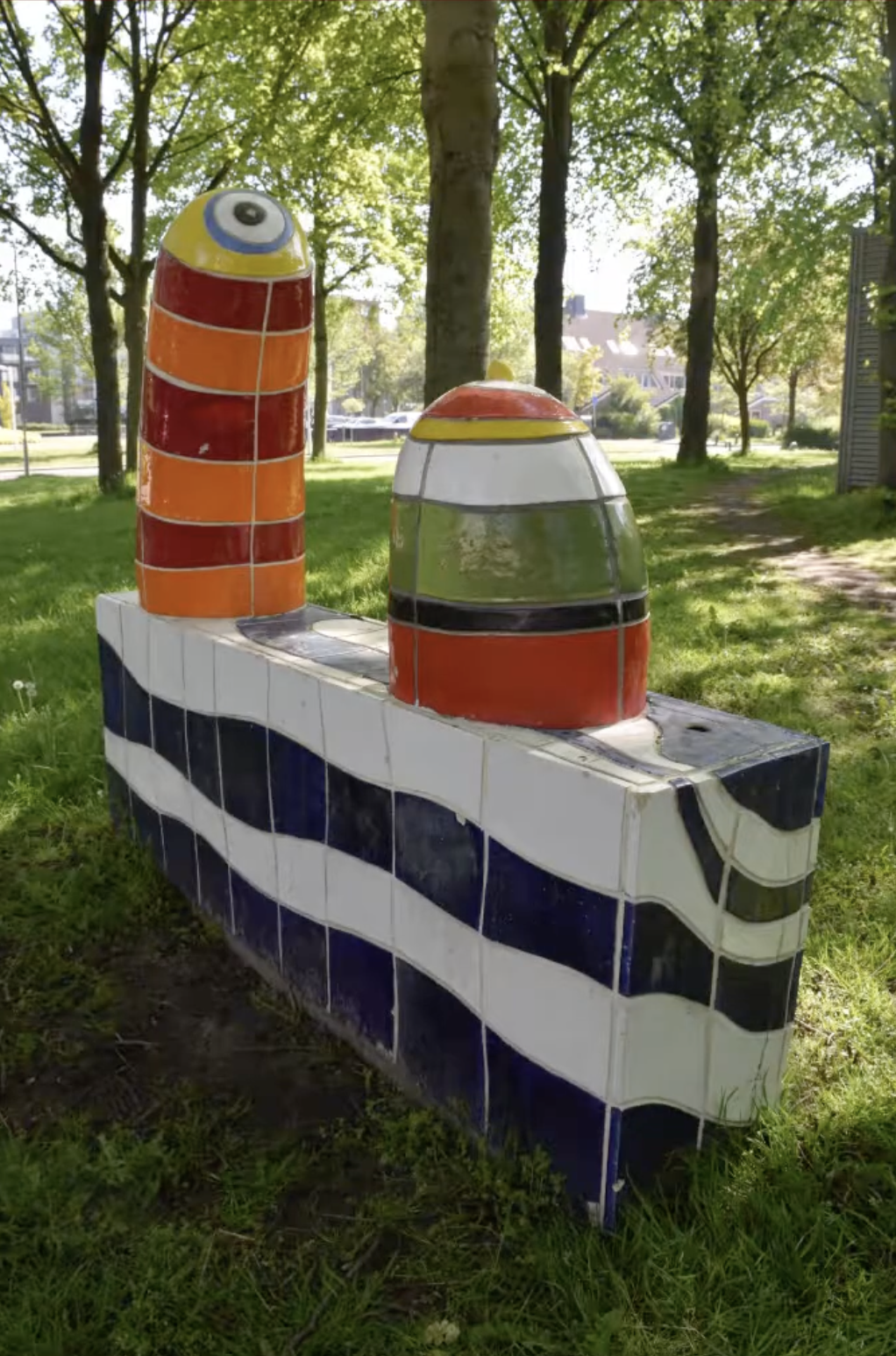 Kleurrijke keramische sculptuur met blauwe en witte golfmotieven op de sokkel en twee felgekleurde torenvormige elementen in rood, oranje en geel, opgesteld in een groenstrook langs een woonstraat.