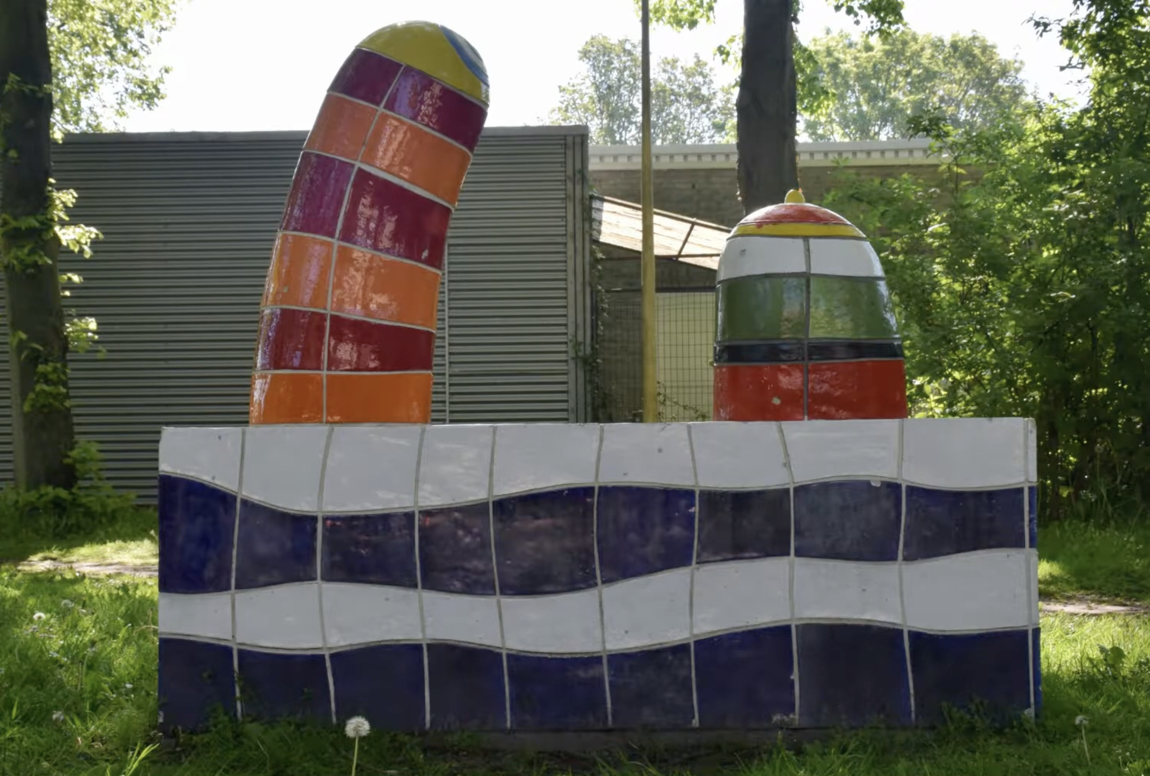 Kleurrijke keramische sculptuur met blauwe en witte golfmotieven op de sokkel en twee felgekleurde torenvormige elementen in rood, oranje en geel, opgesteld in een groenstrook langs een woonstraat.