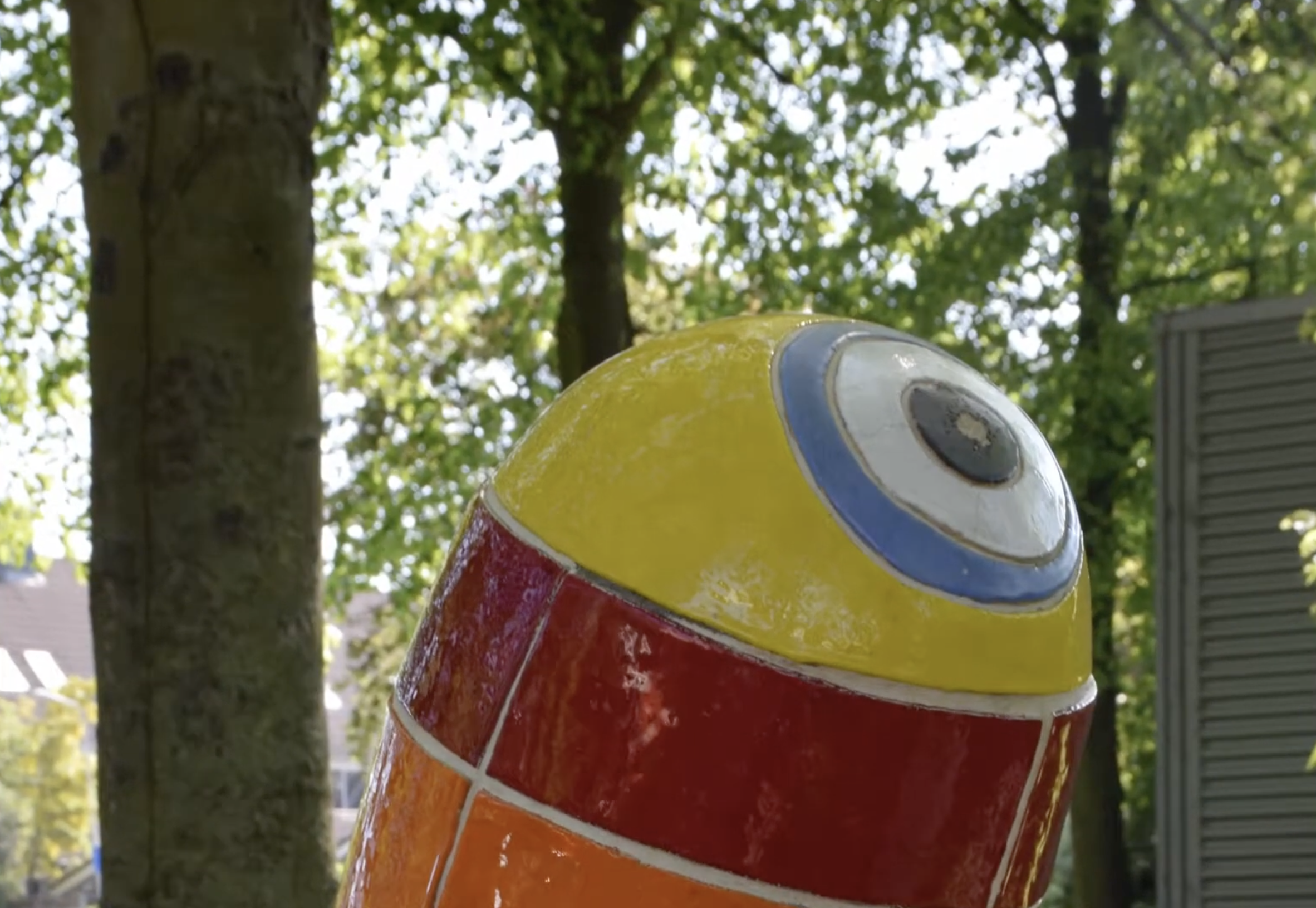 Kleurrijke keramische sculptuur met blauwe en witte golfmotieven op de sokkel en twee felgekleurde torenvormige elementen in rood, oranje en geel, opgesteld in een groenstrook langs een woonstraat.