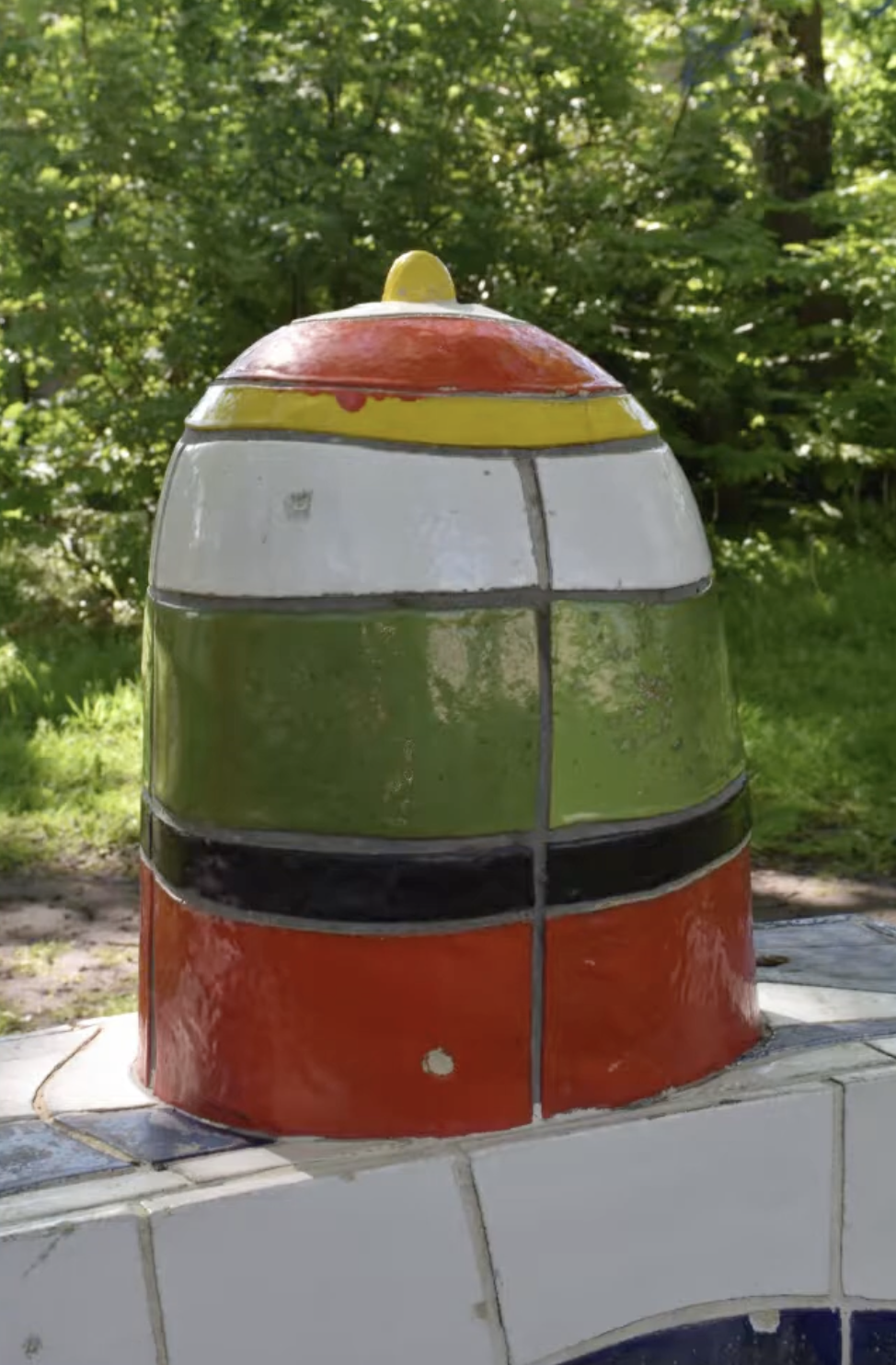 Kleurrijke keramische sculptuur met blauwe en witte golfmotieven op de sokkel en twee felgekleurde torenvormige elementen in rood, oranje en geel, opgesteld in een groenstrook langs een woonstraat.