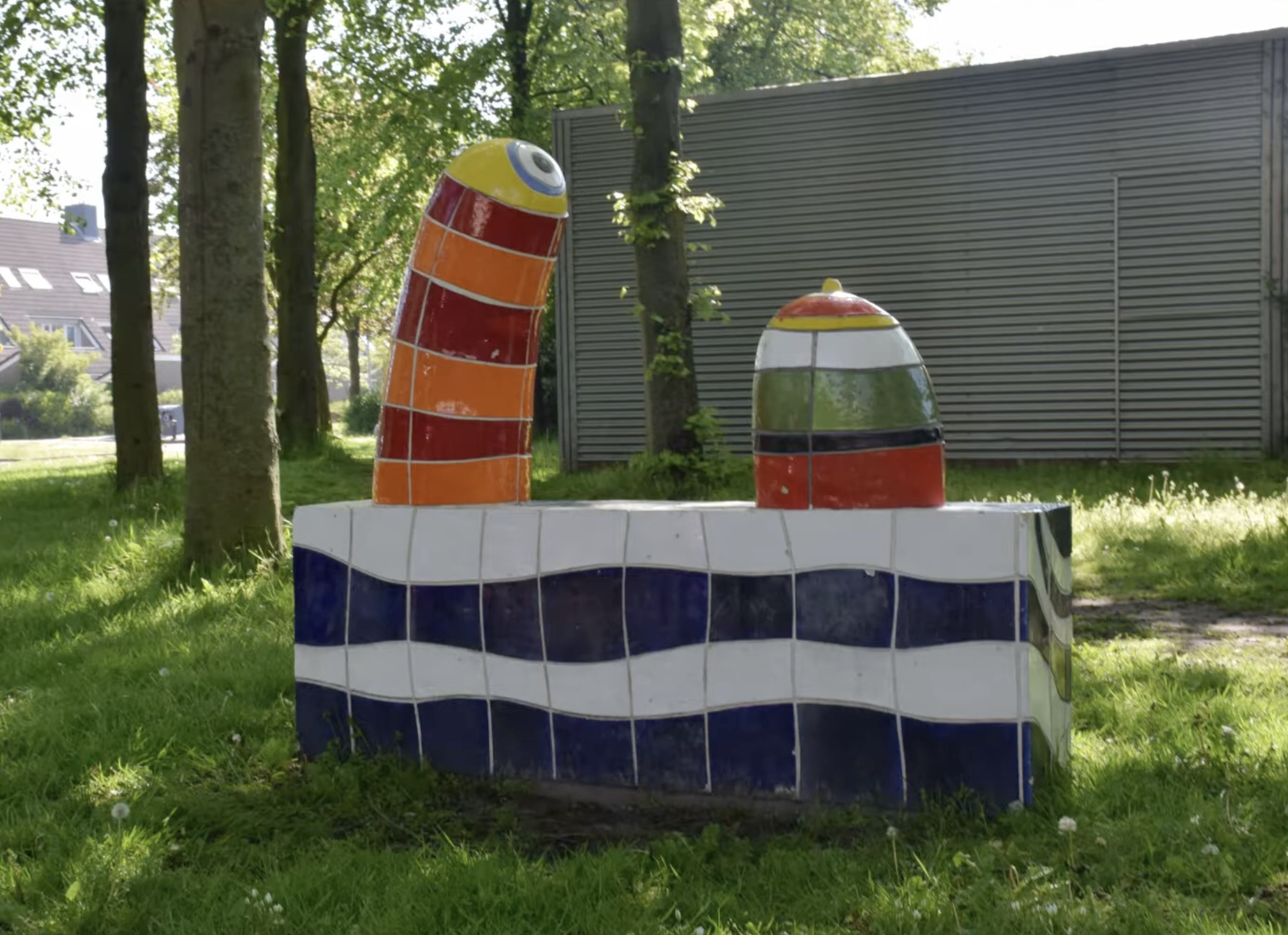 Kleurrijke keramische sculptuur met blauwe en witte golfmotieven op de sokkel en twee felgekleurde torenvormige elementen in rood, oranje en geel, opgesteld in een groenstrook langs een woonstraat.