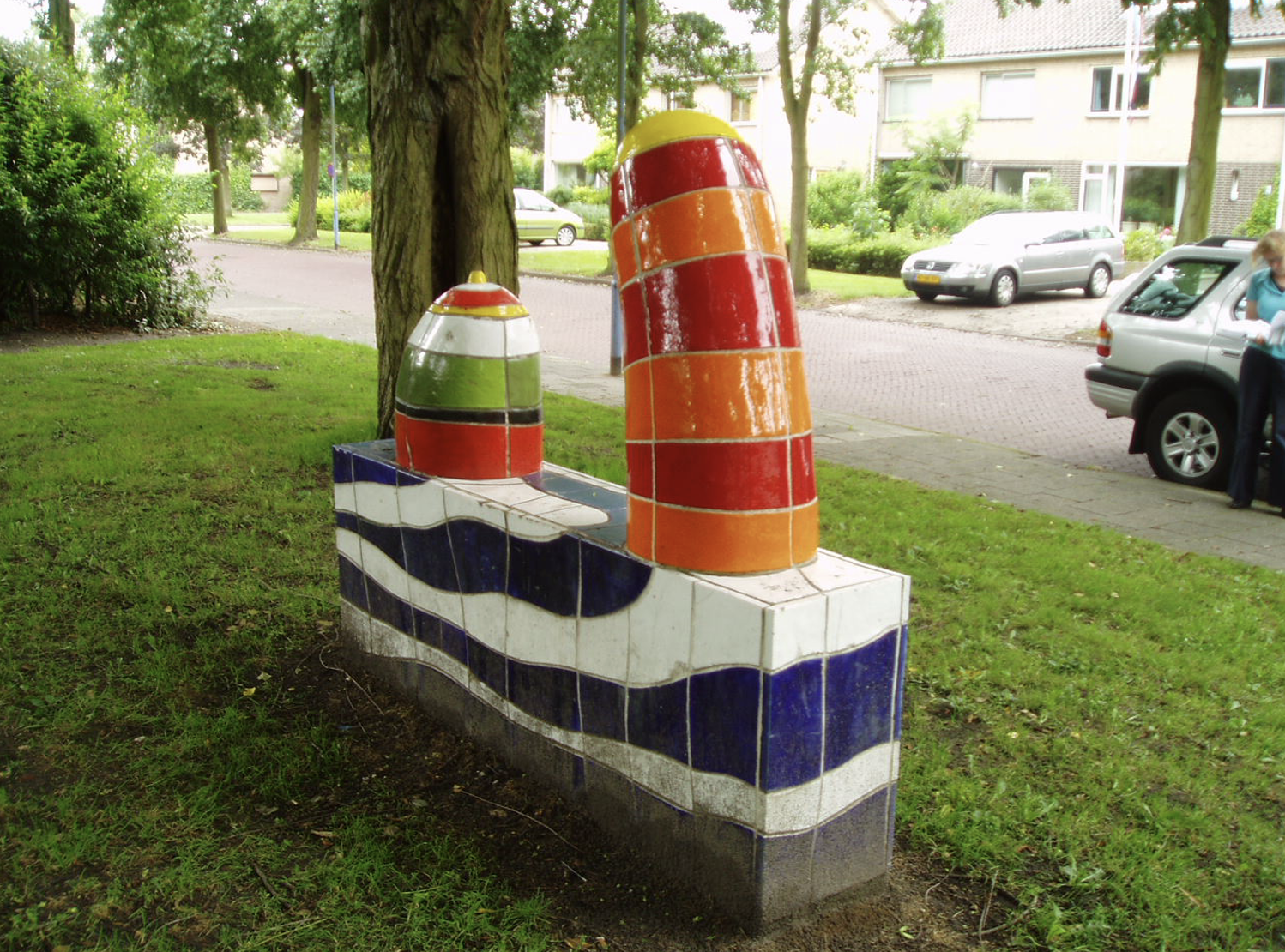Kleurrijke keramische sculptuur met blauwe en witte golfmotieven op de sokkel en twee felgekleurde torenvormige elementen in rood, oranje en geel, opgesteld in een groenstrook langs een woonstraat.