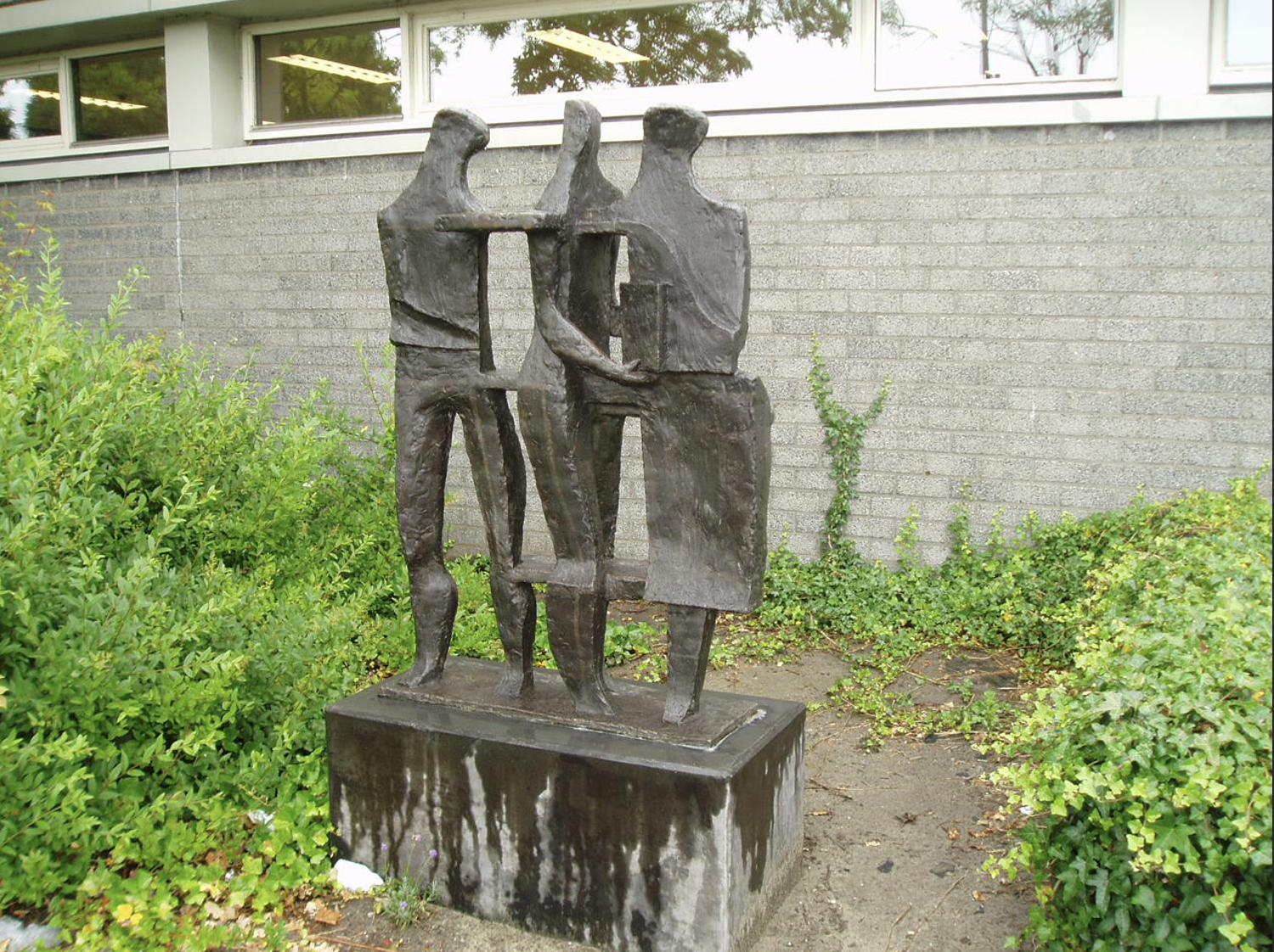 Bronzen sculptuur van drie gestileerde menselijke figuren die samen iets vasthouden, op een betonnen sokkel voor een grijze bakstenen muur.