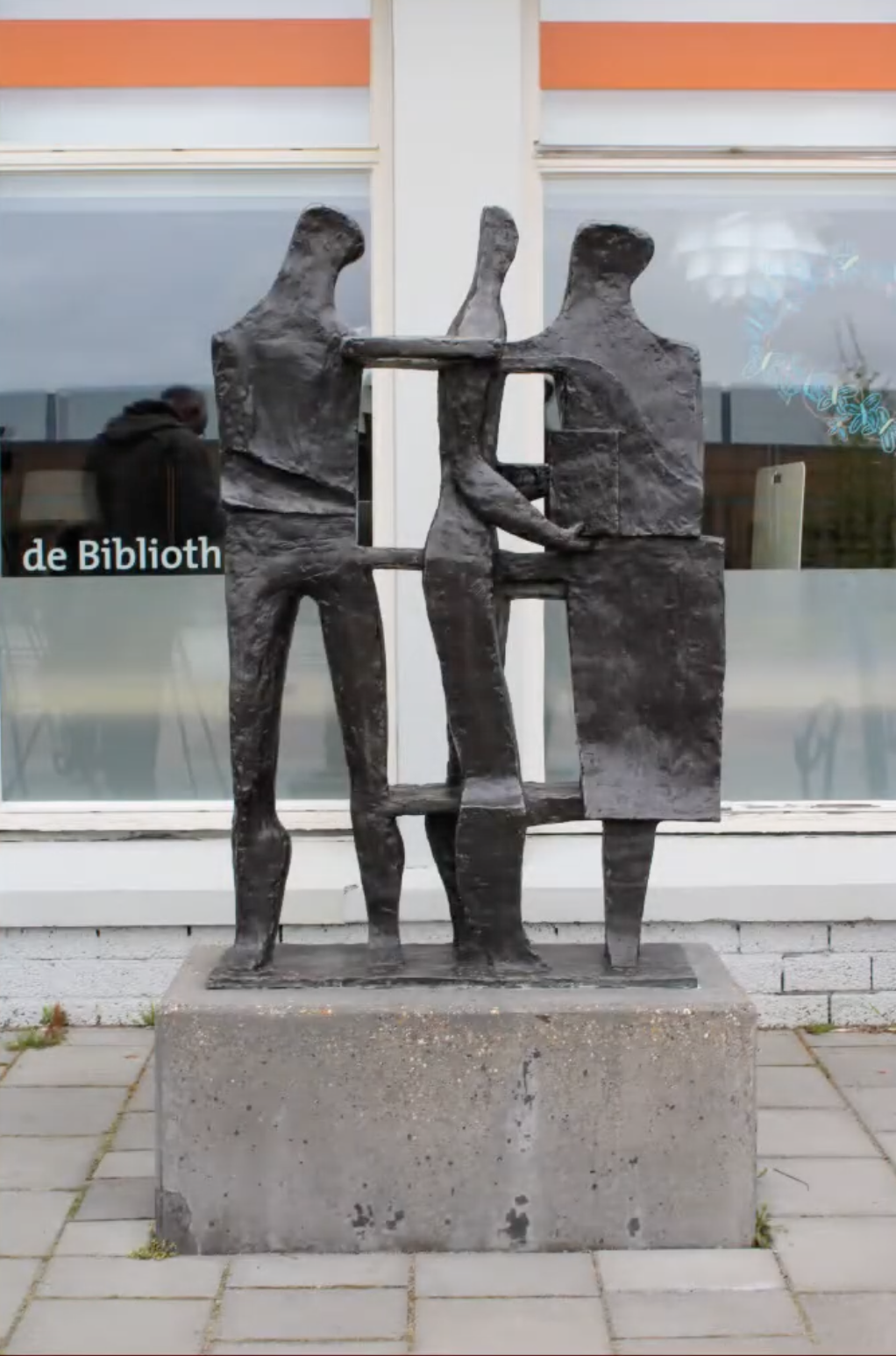 Bronzen sculptuur van drie gestileerde menselijke figuren die samen iets vasthouden, op een betonnen sokkel voor een grijze bakstenen muur.